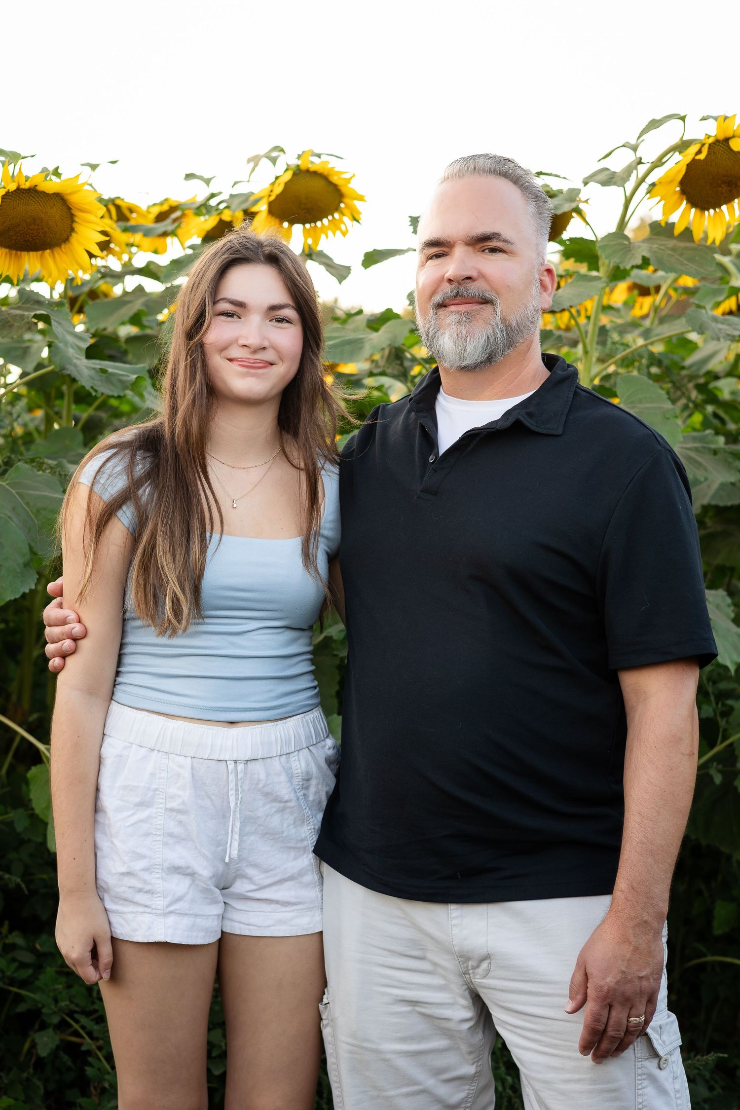 Two people pose together in casual attire against a backdrop of towering sunflowers.