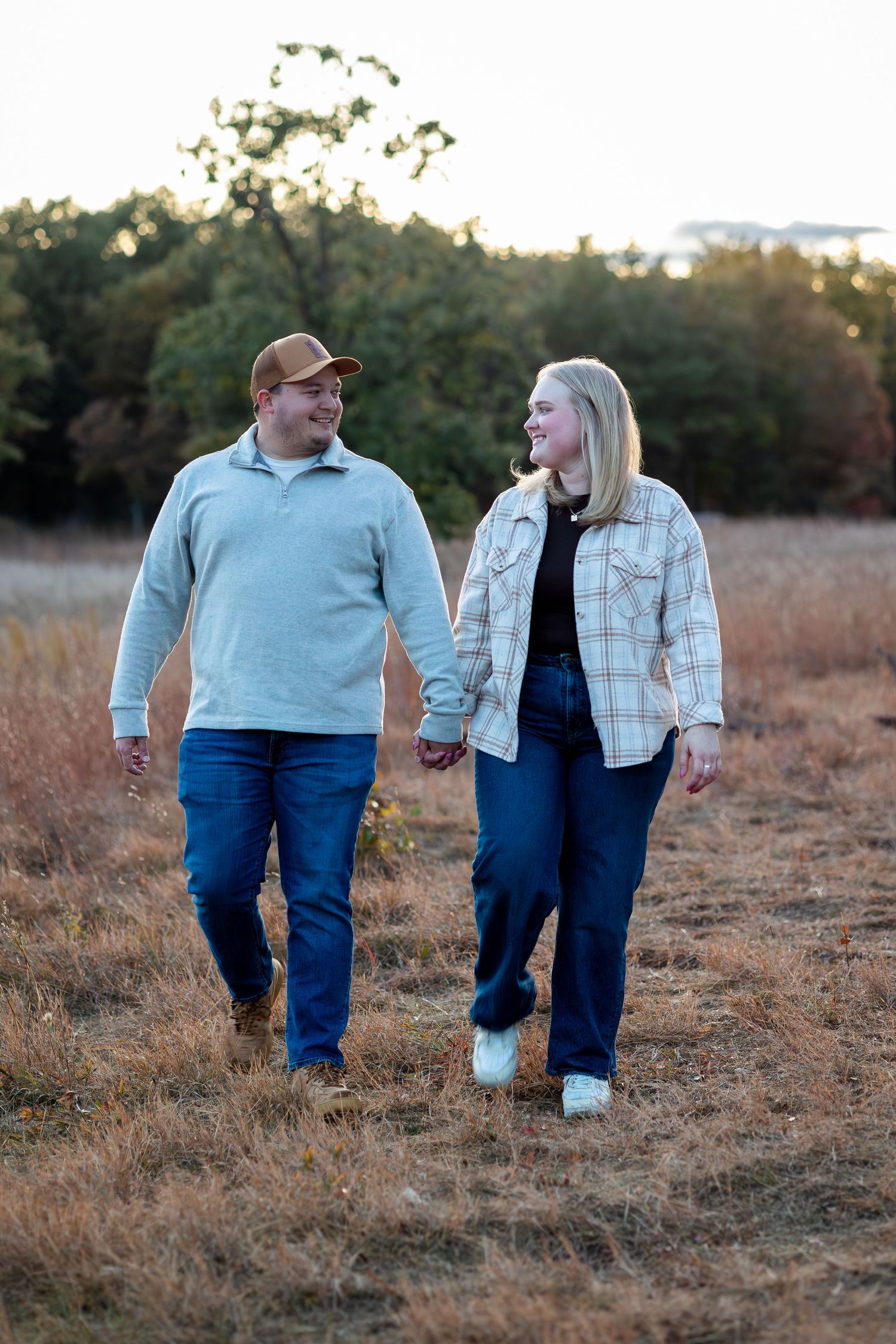 A couple walks hand in hand through a golden field at sunset wearing coordinating casual outfits.
