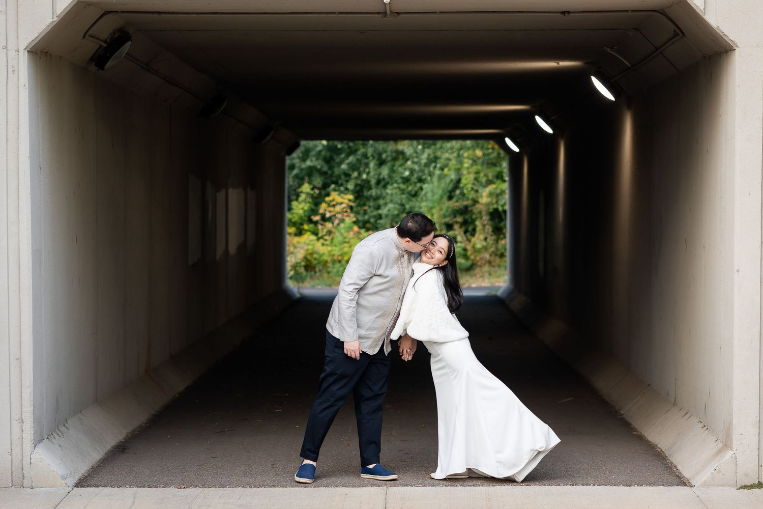 A couple shares a playful moment in a concrete tunnel with natural light at the end.