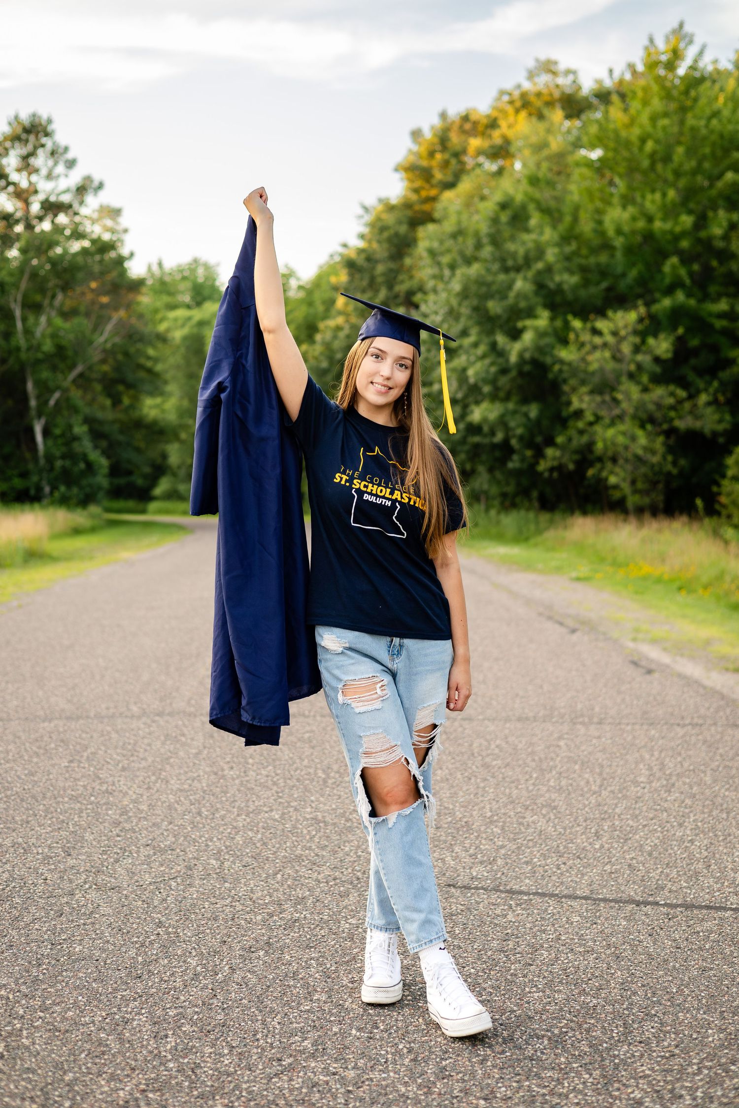 Graduate in school t-shirt and ripped jeans poses with cap on outdoor path.