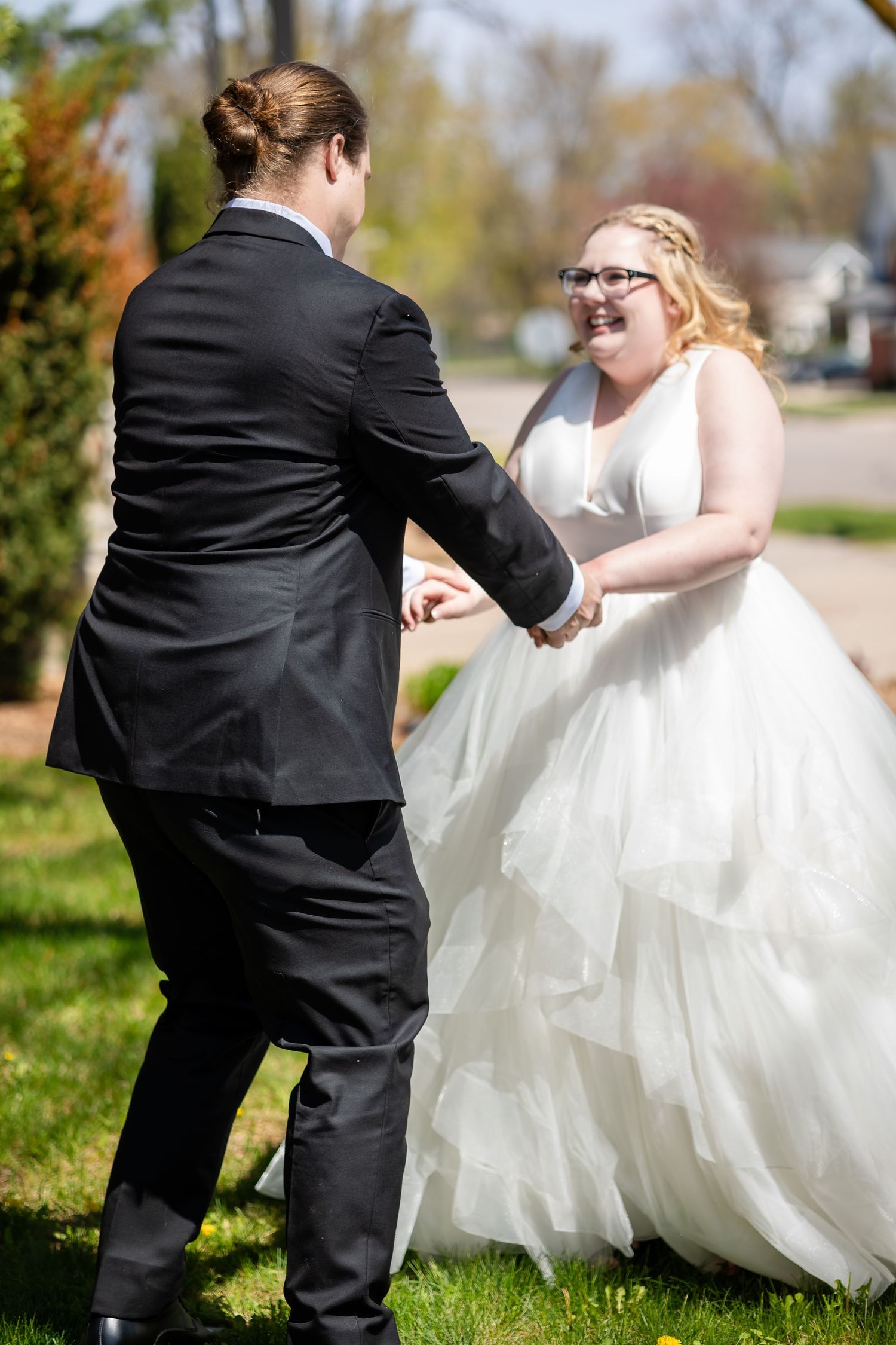Bride in white tulle gown laughing while holding hands with groom in black suit outdoors on sunny wedding day.