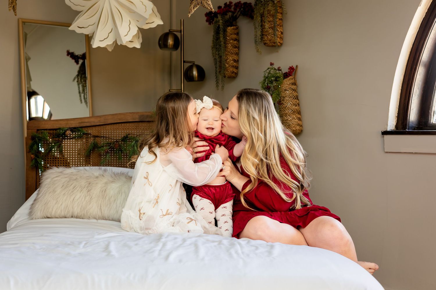 A mother and young daughter kiss a baby girl in a red dress while sitting on a white bed in a cozy decorated room.