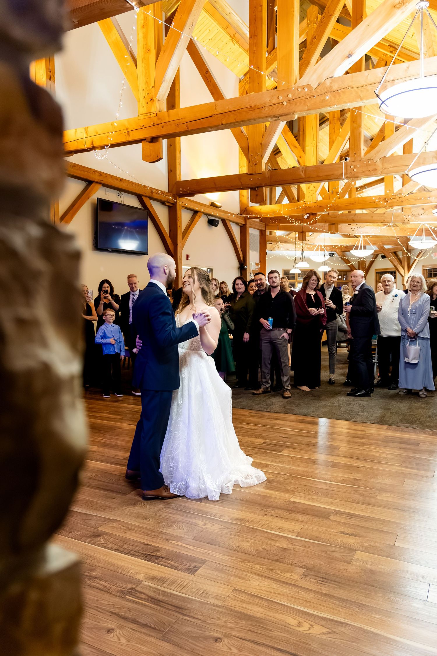 Bride and groom share their first dance in a rustic wooden venue while wedding guests watch in the background.