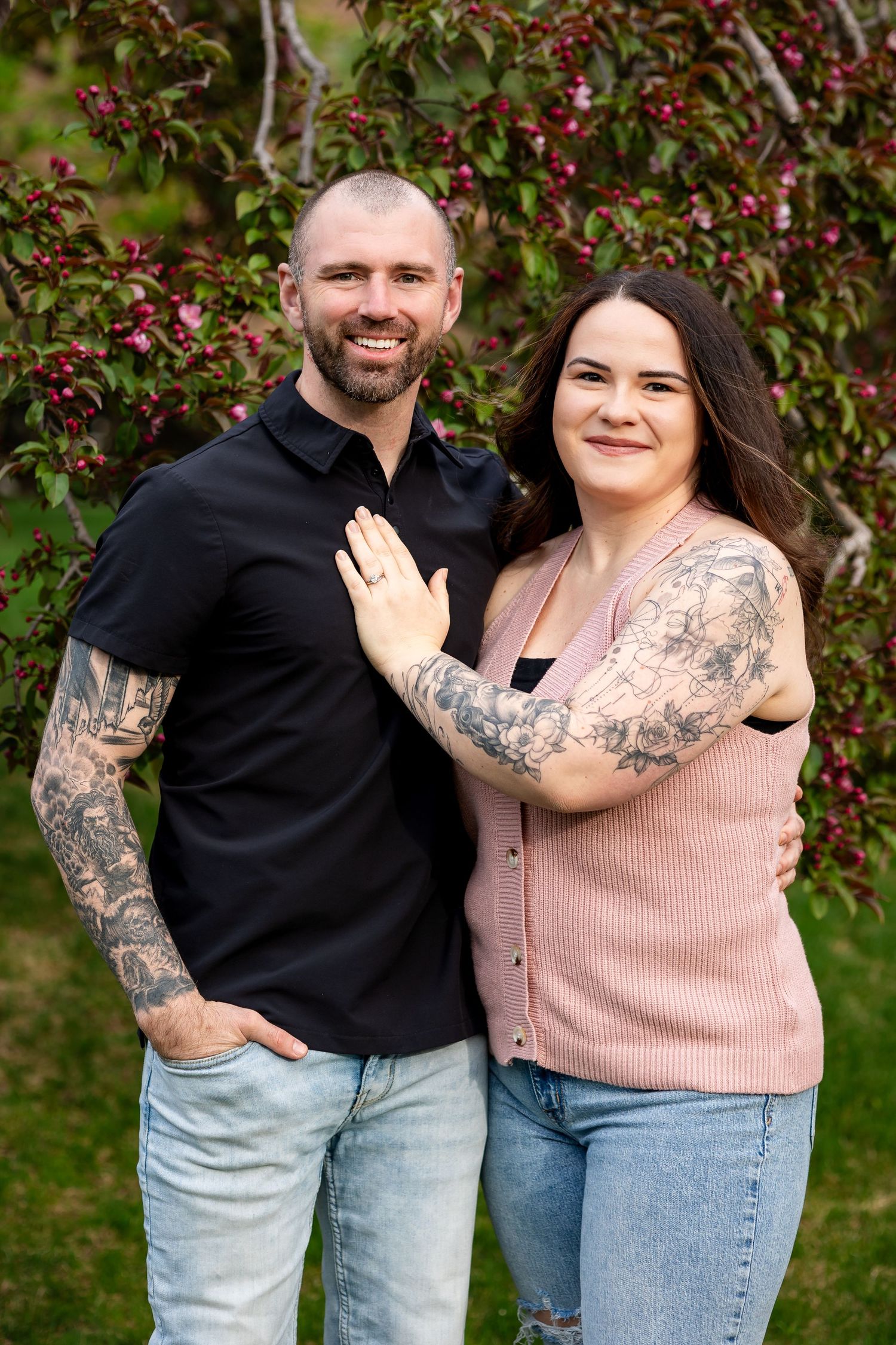 Smiling couple with tattoos posing outdoors near flowering tree, woman showing engagement ring on man's chest.