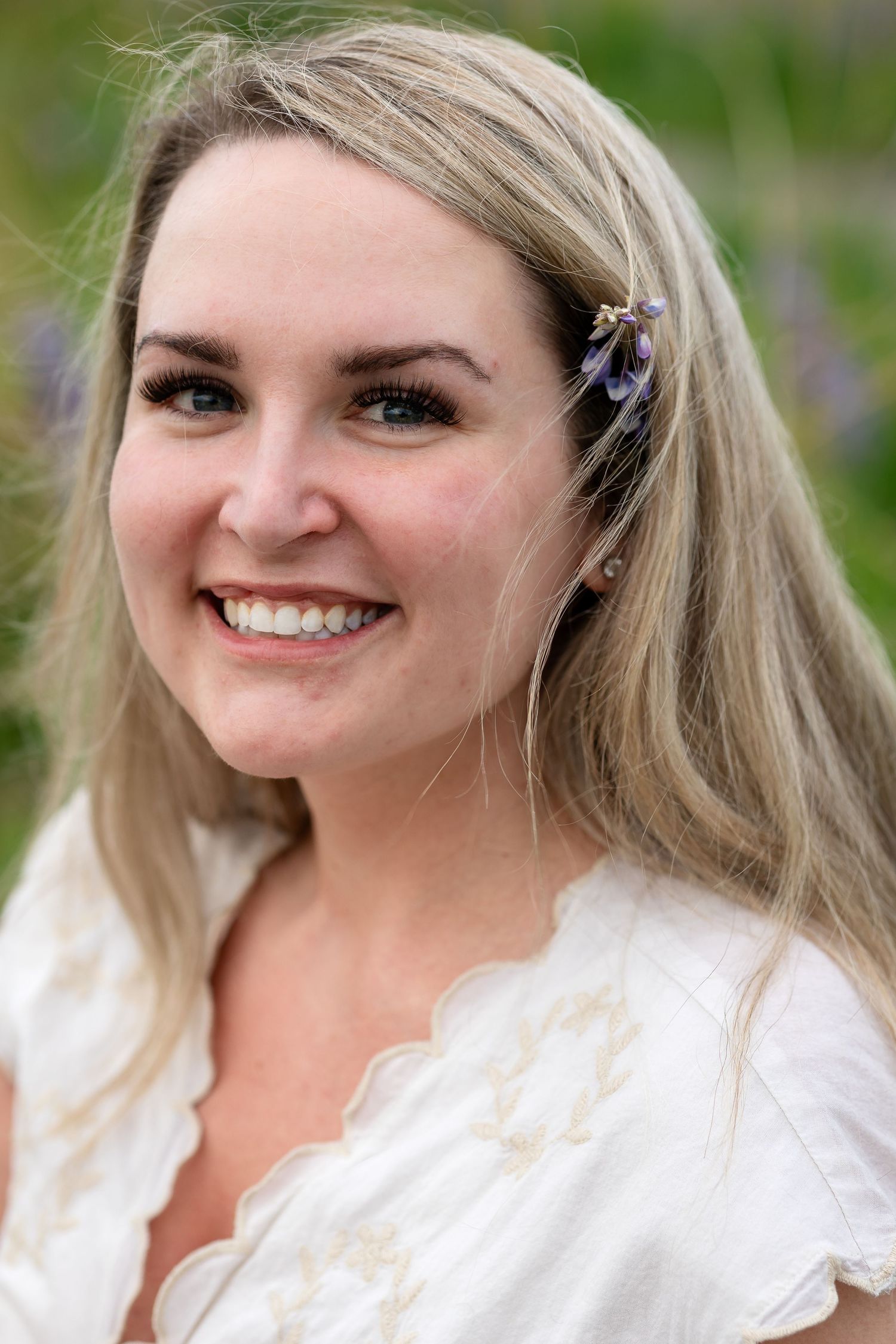Smiling blonde woman with purple flower hair clip wearing white dress outdoors in garden setting.