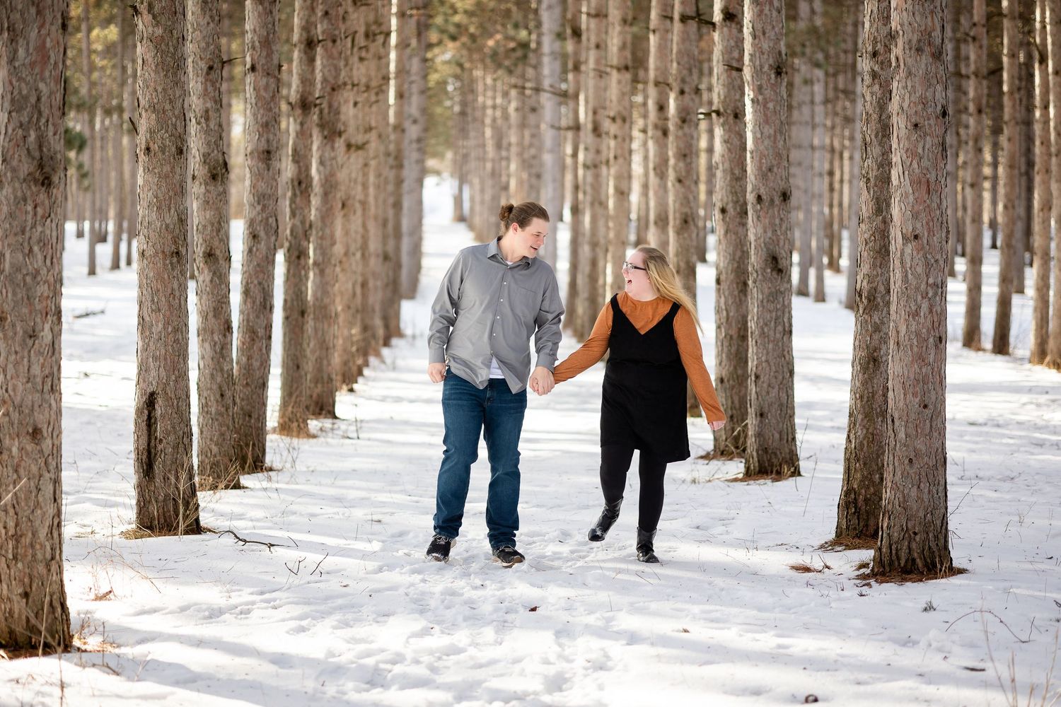 Couple holding hands walking through snow-covered pine tree path in winter, wearing casual cold weather outfits.