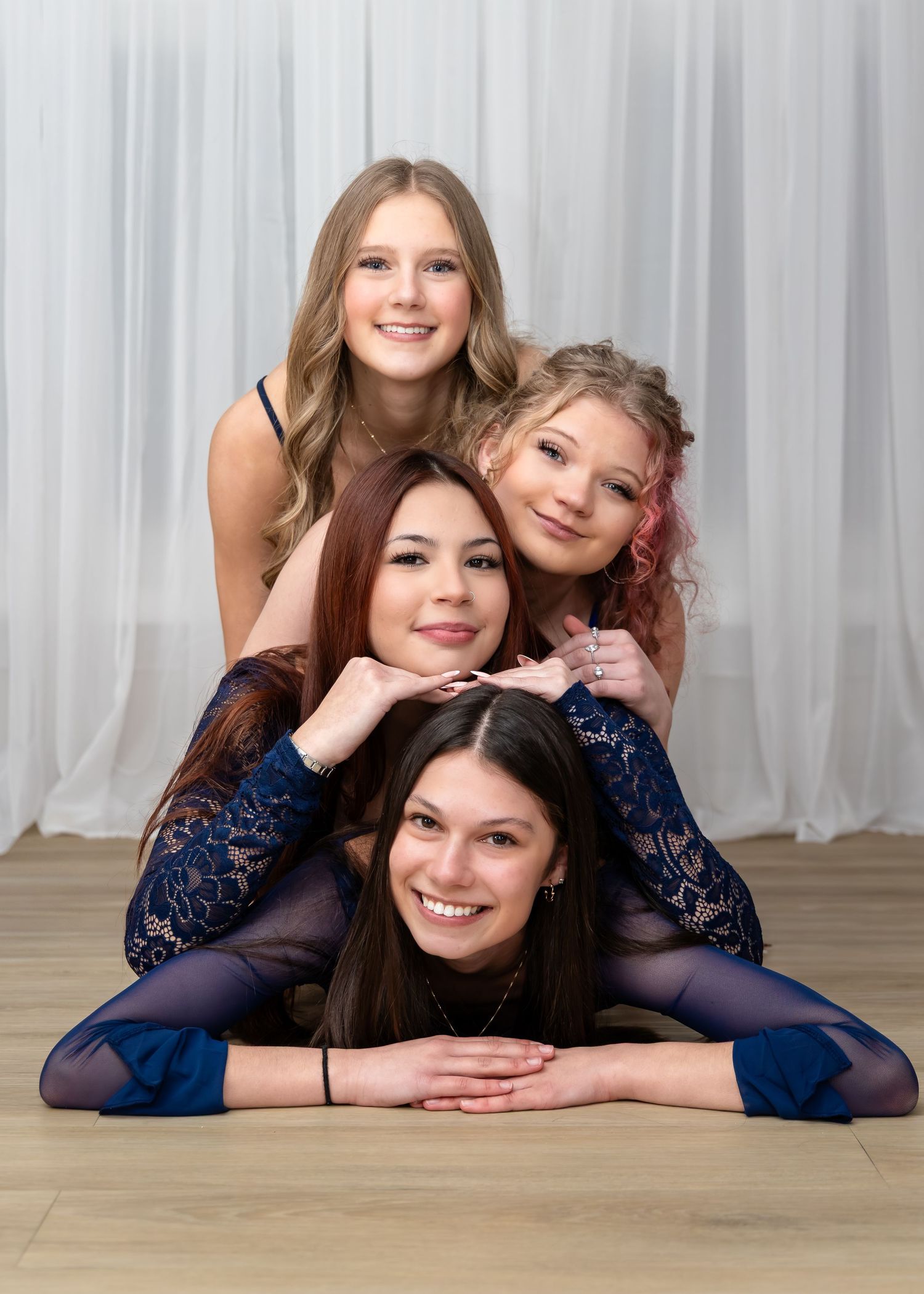 Four young women pose in a stacked pyramid formation, wearing blue outfits, smiling in a bright studio setting.