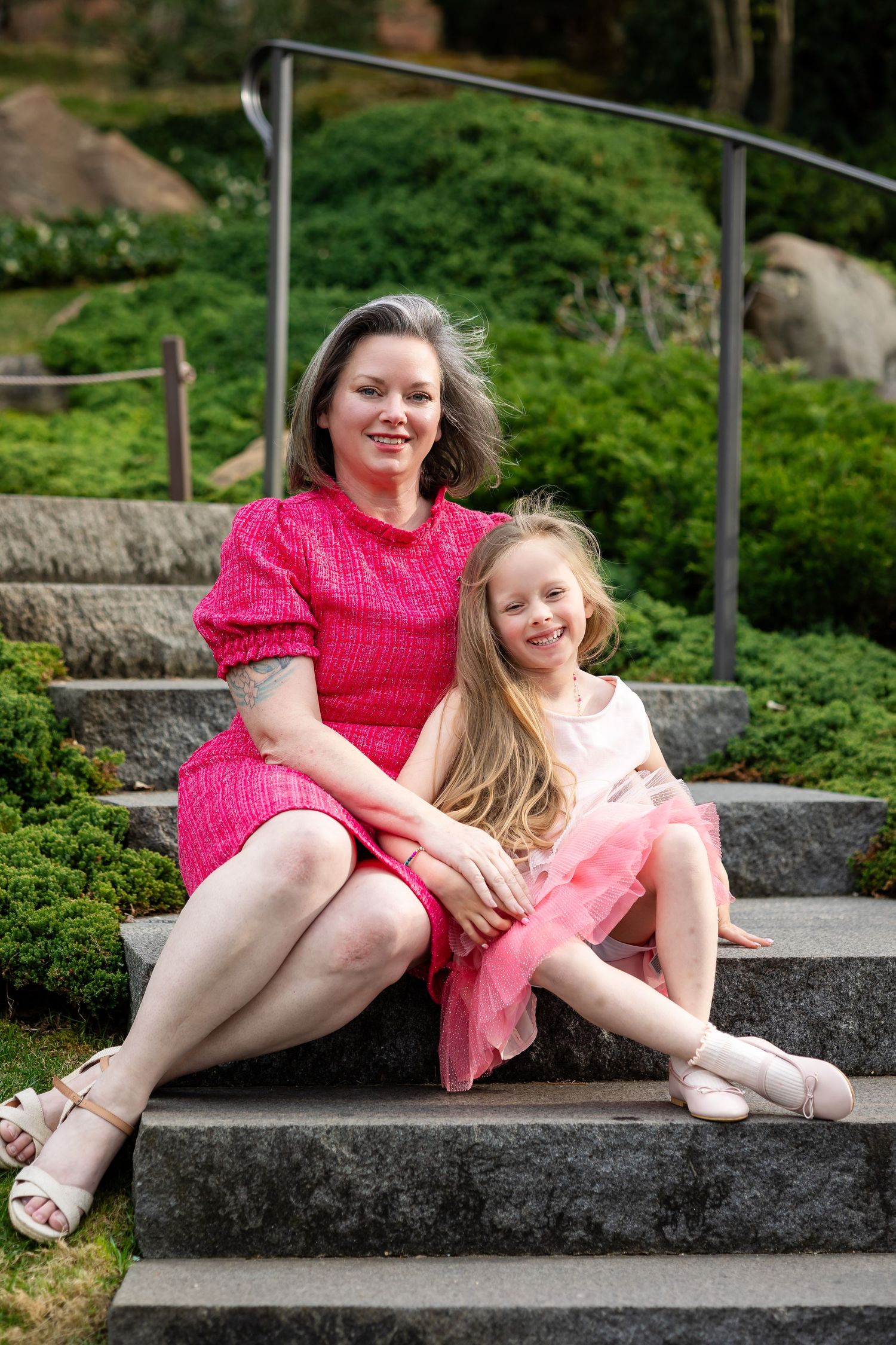 A woman in a pink dress and young girl in a pink skirt smile while sitting together on stone steps in a garden.