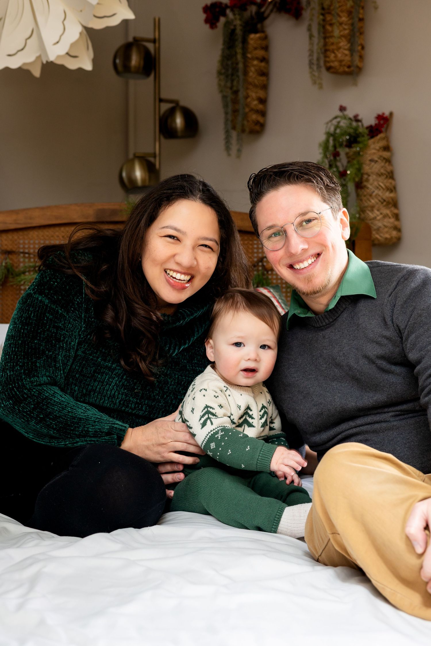 Happy family portrait with two smiling parents and their baby sitting on a bed with festive holiday decor.