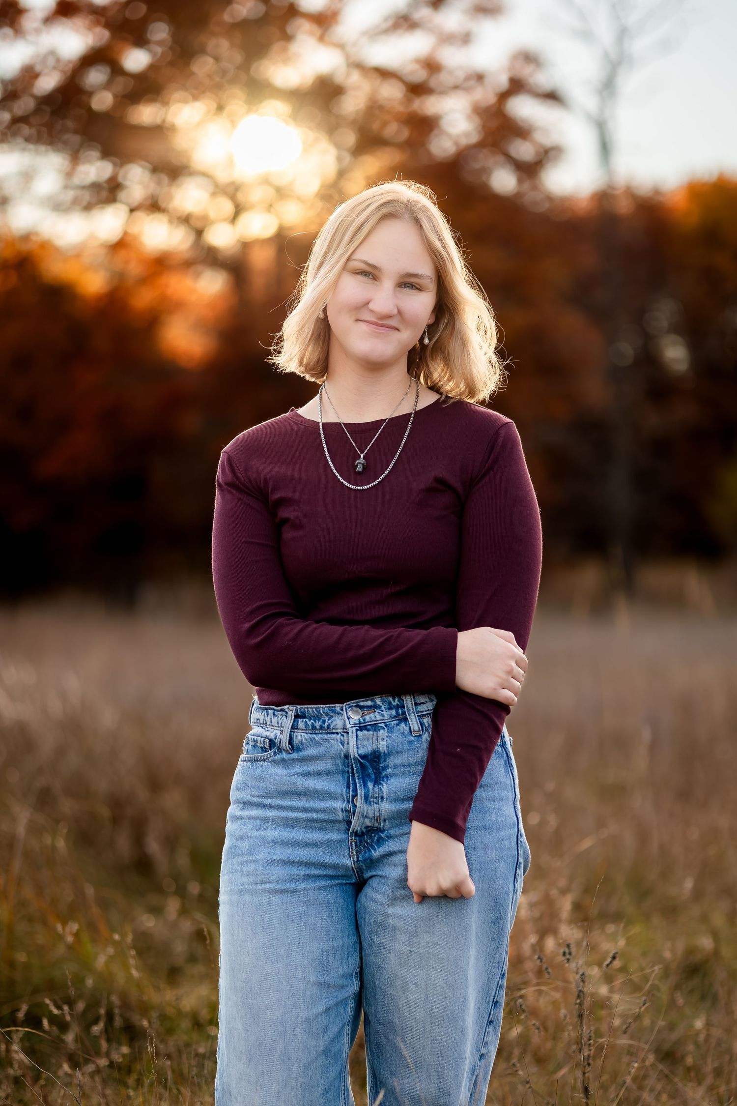 A person in a burgundy sweater and jeans poses outdoors during golden hour sunset in a field of tall grass.