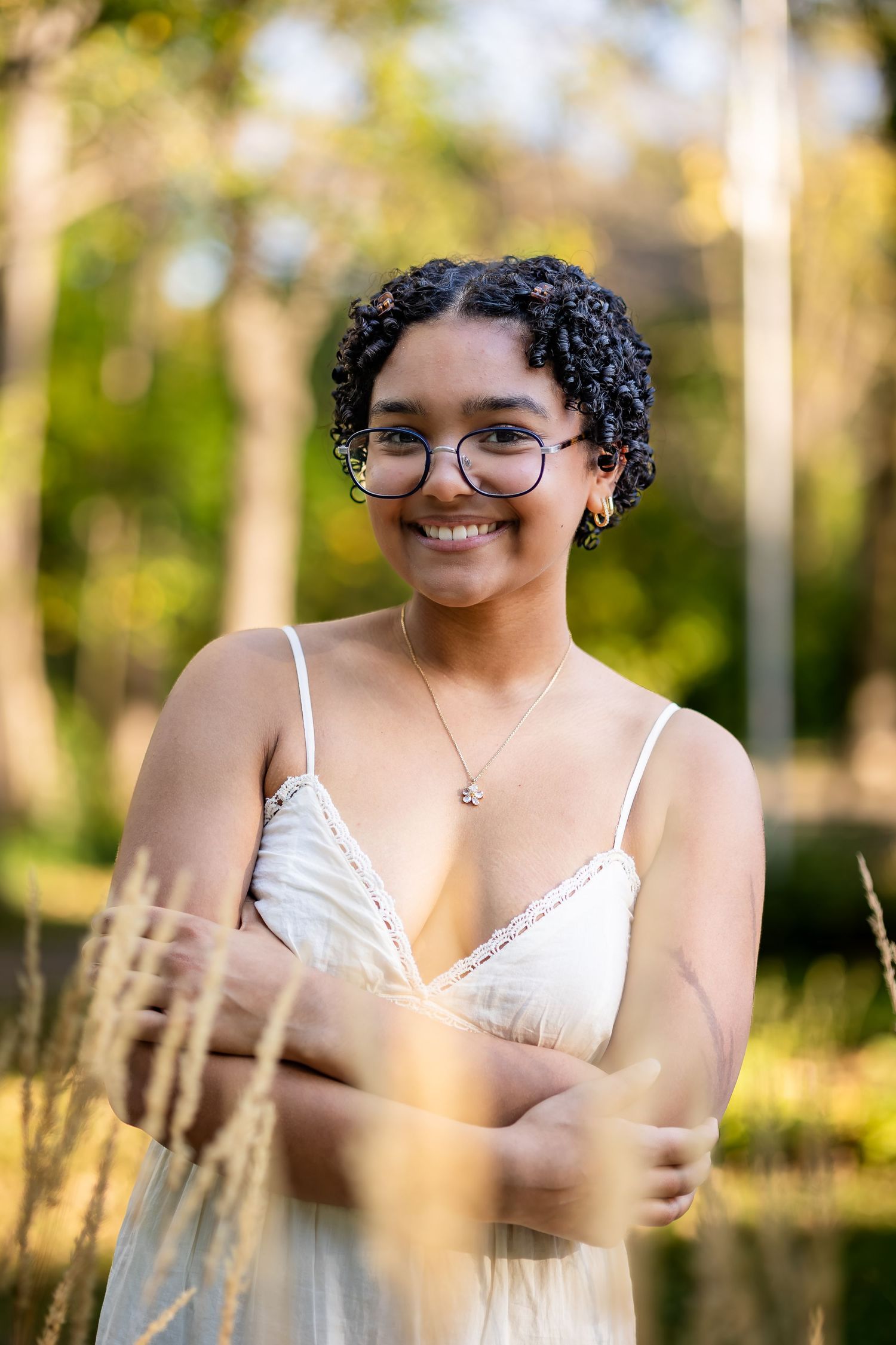 Outdoor portrait with sunlit trees and foliage in background, wearing glasses and white lace top.