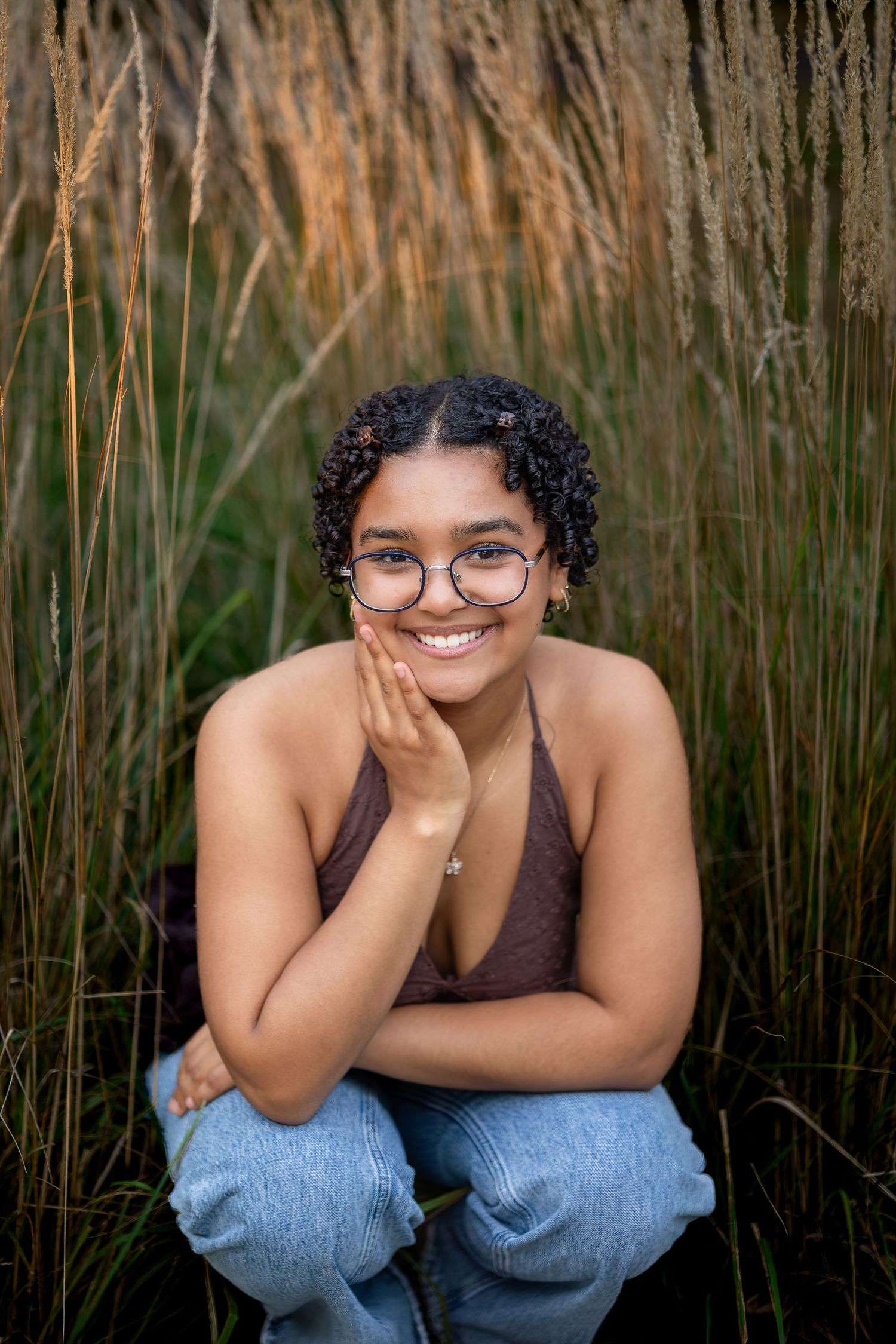 Person in brown top and jeans poses with hand on chin among tall ornamental grasses at sunset.