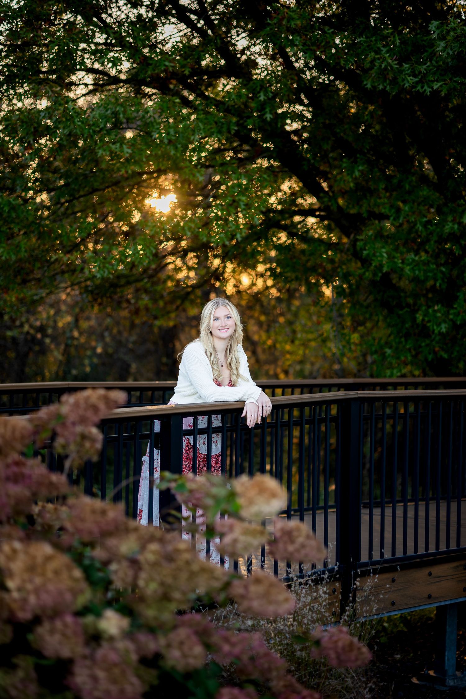 Woman in white sweater leans on dark railing with autumn foliage in background.