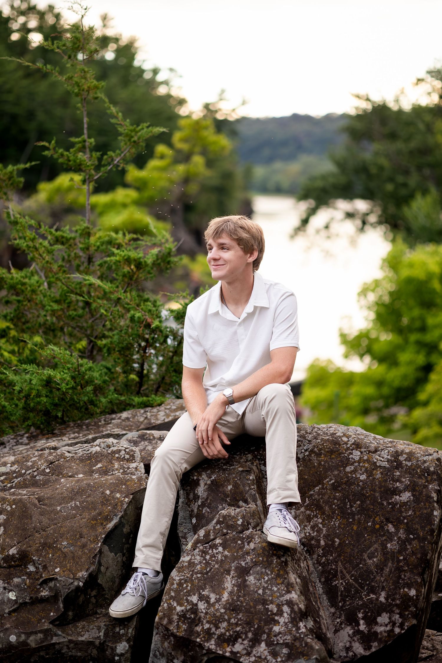 Person in white shirt sits casually on large rock formation overlooking water during sunset.