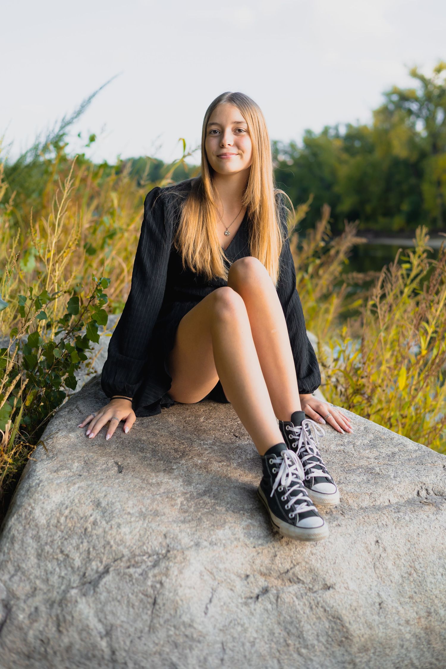 Person sitting on large rock wearing black top and sneakers near waterfront.
