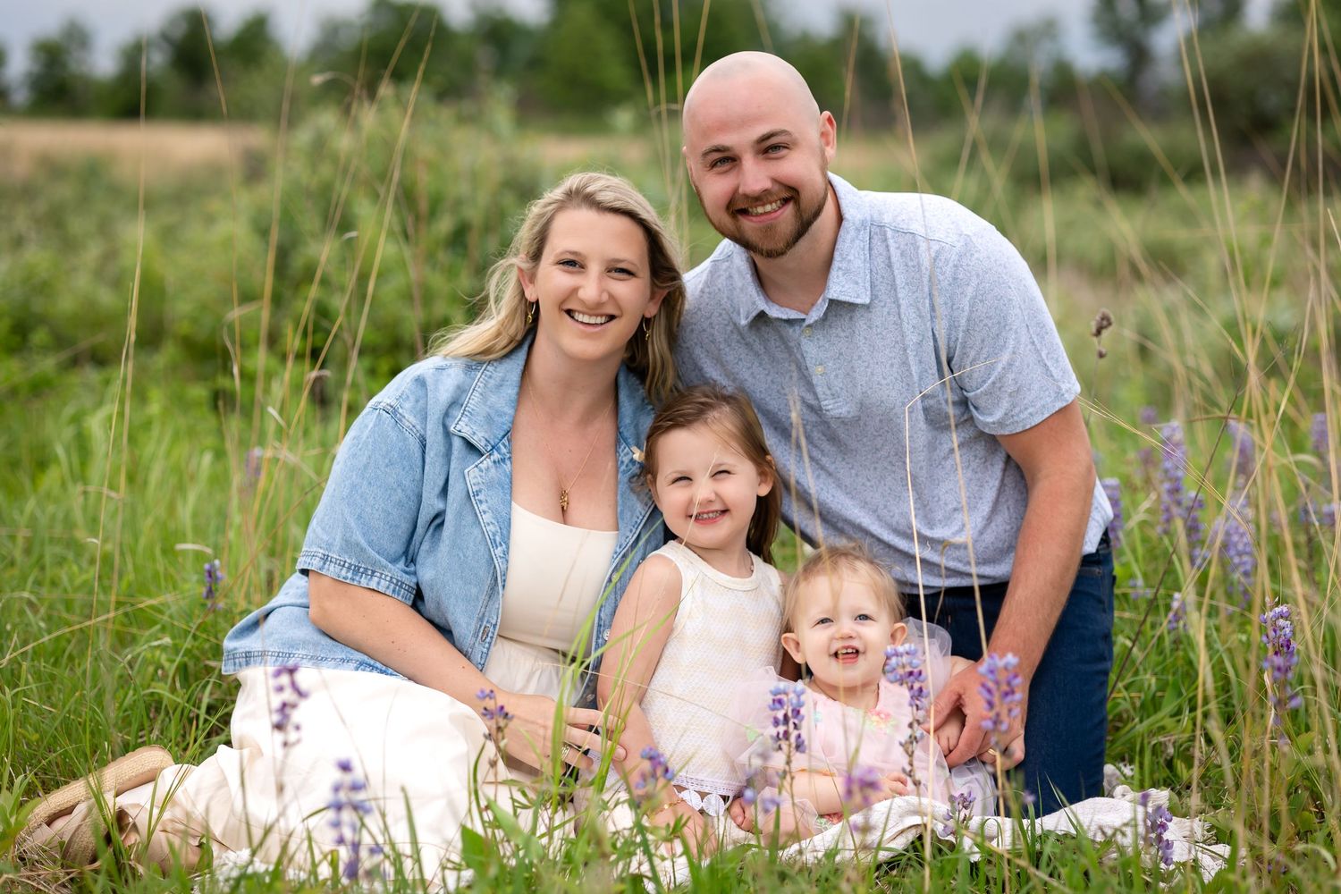 Family of four smiling outdoors in a green meadow with wildflowers, parents and two young daughters seated together.