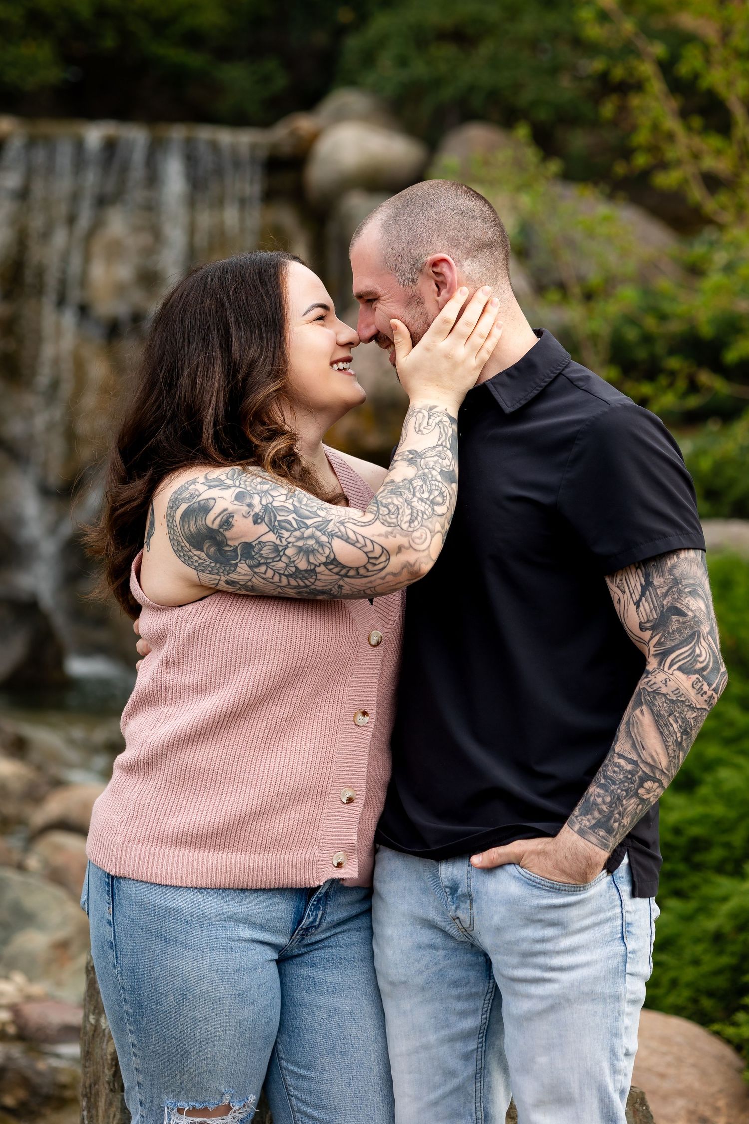 A tattooed couple shares a romantic moment near a waterfall, the woman smiling as she gently cups the mans face.