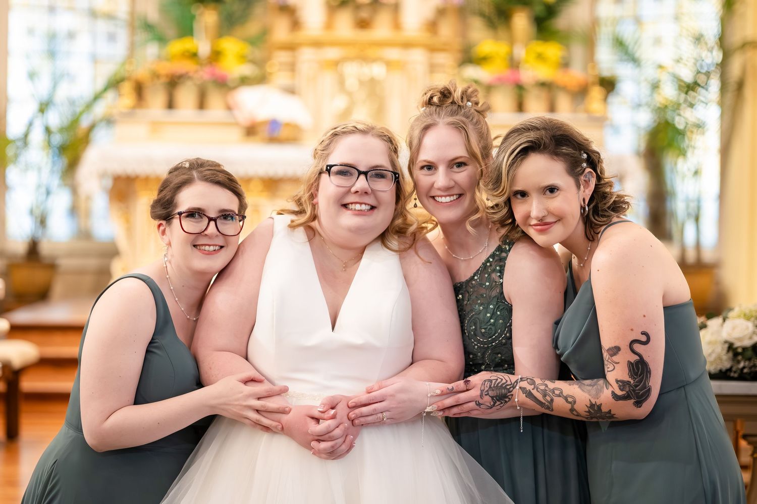 Smiling bride with three bridesmaids in sage green dresses posing together in front of church altar.