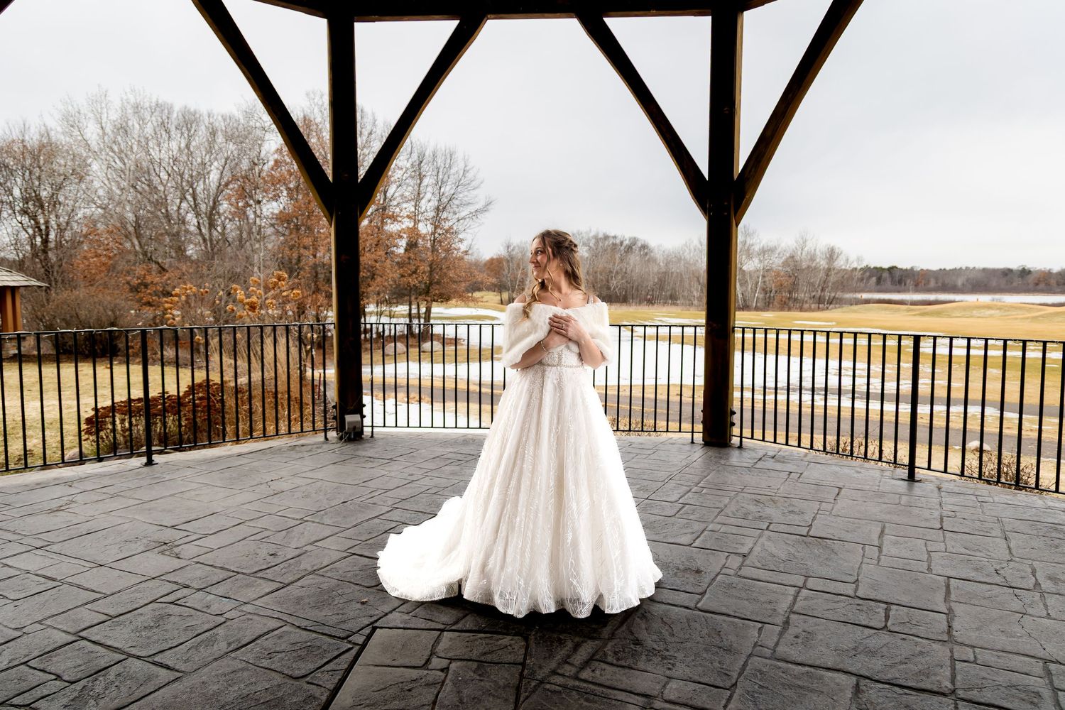 Bride in elegant white off-shoulder ball gown twirling on a covered pavilion overlooking a serene lake in autumn.