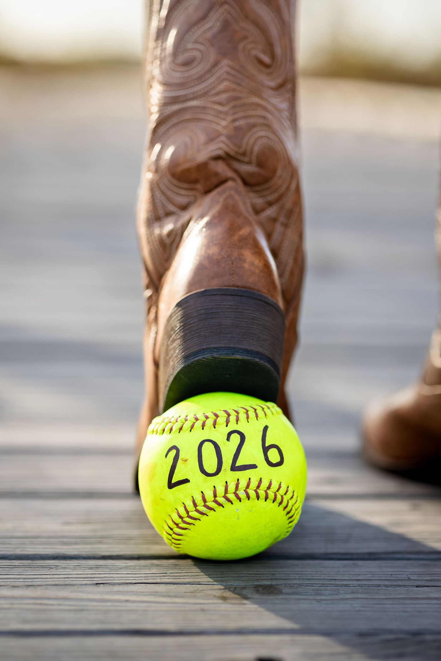 Brown leather cowboy boot steps on a bright yellow softball against a wooden deck surface.