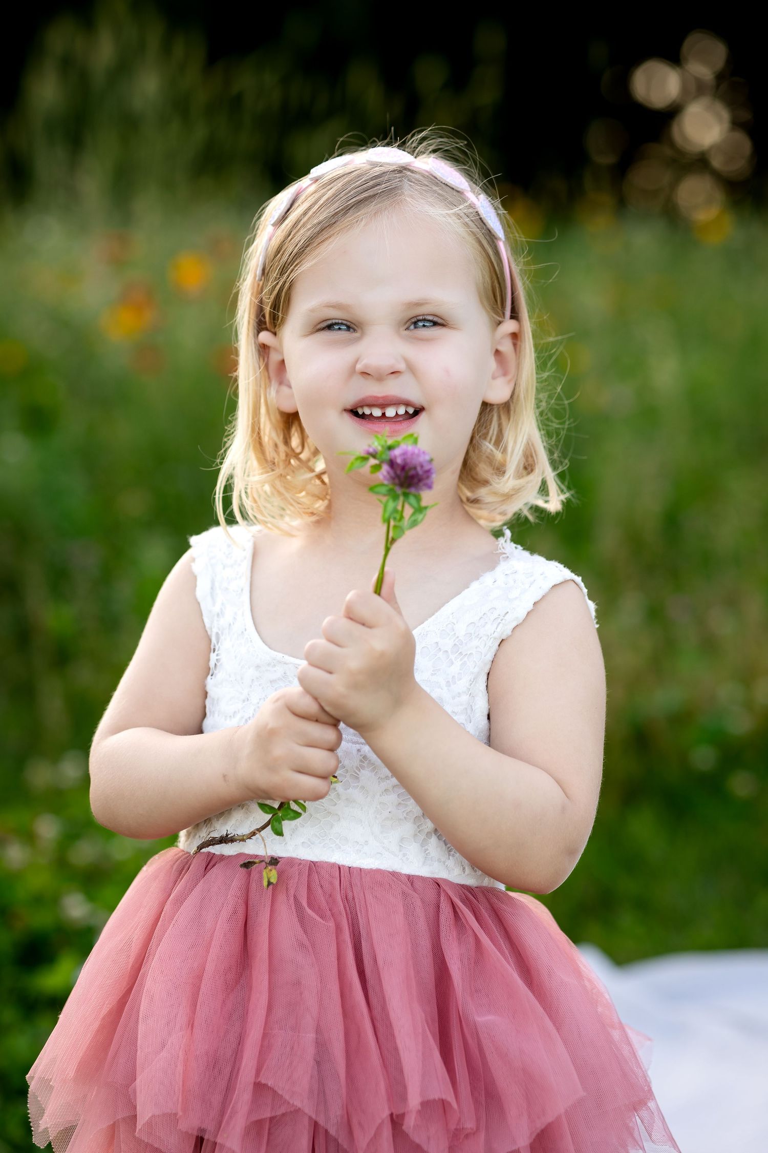 Young child in pink tulle dress poses playfully with flowers in a garden setting.