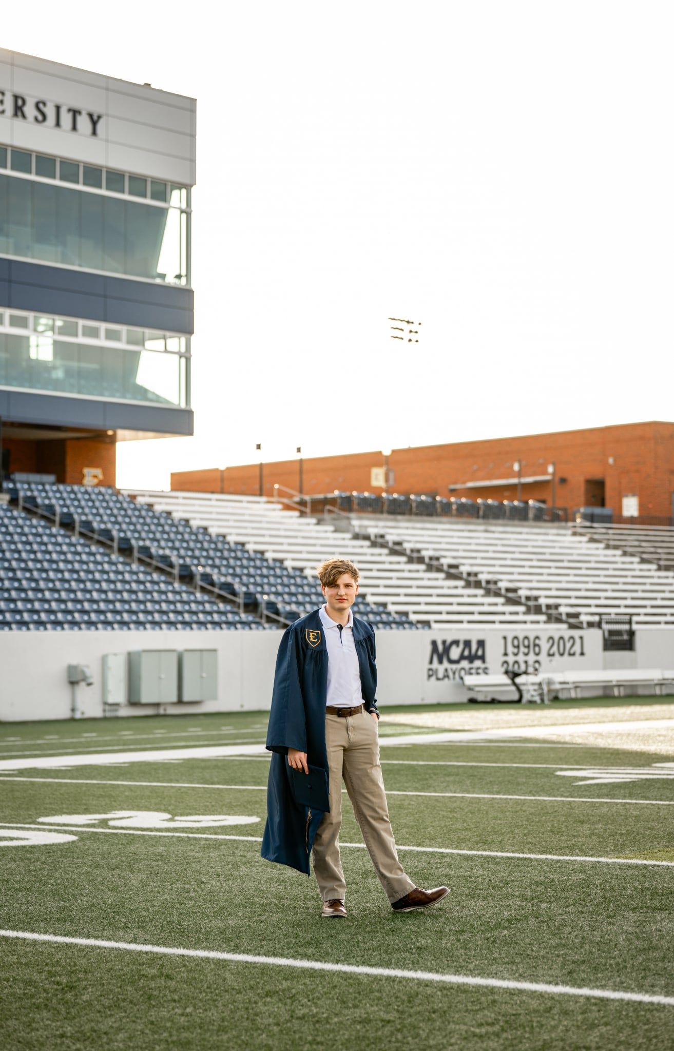 Graduate in cap and gown walks across football field with empty stadium bleachers and university building in background.
