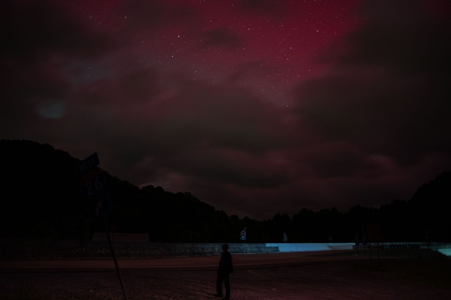 Silhouetted figure stands on beach under dark red night sky with glowing blue horizon.