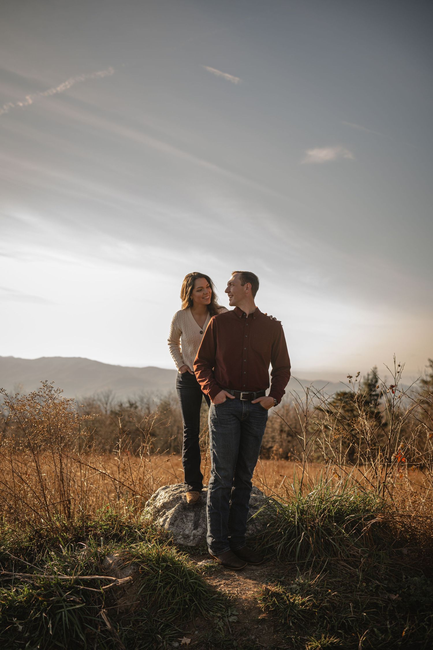 A couple stands together on a mountaintop during golden hour with scenic mountain views in the background.