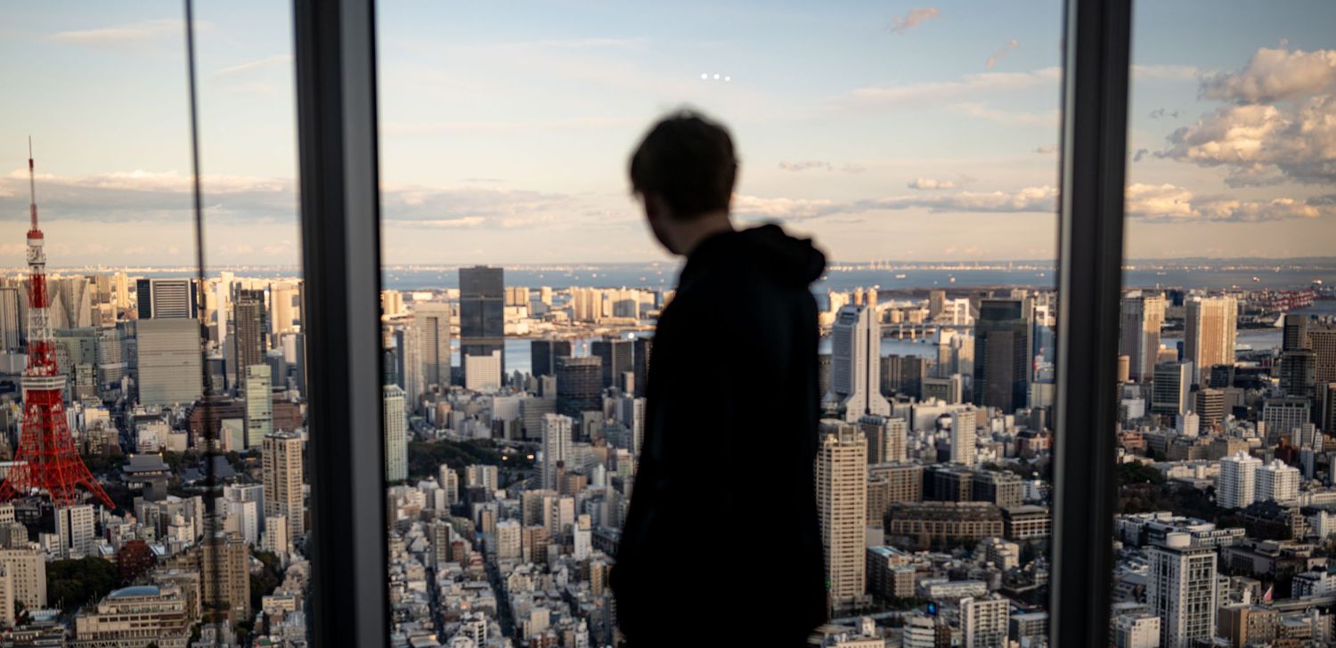 Silhouette of person looking out over Tokyo cityscape through large window during sunset.