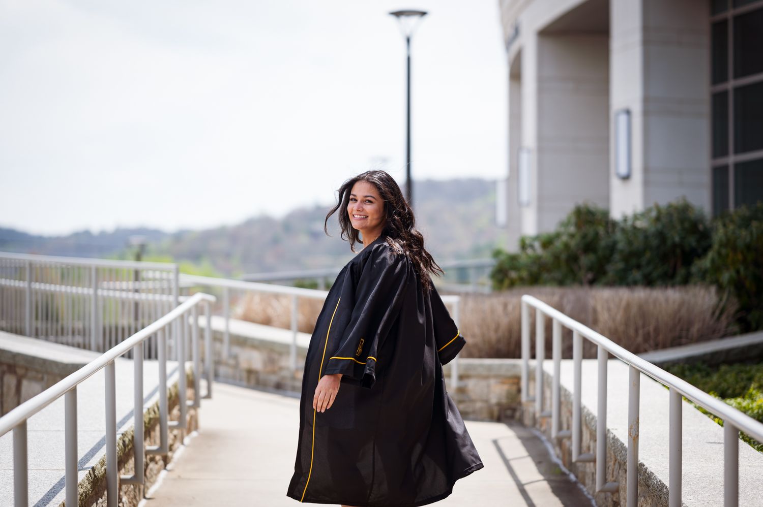 A graduate in black cap and gown poses confidently on a walkway with railings.