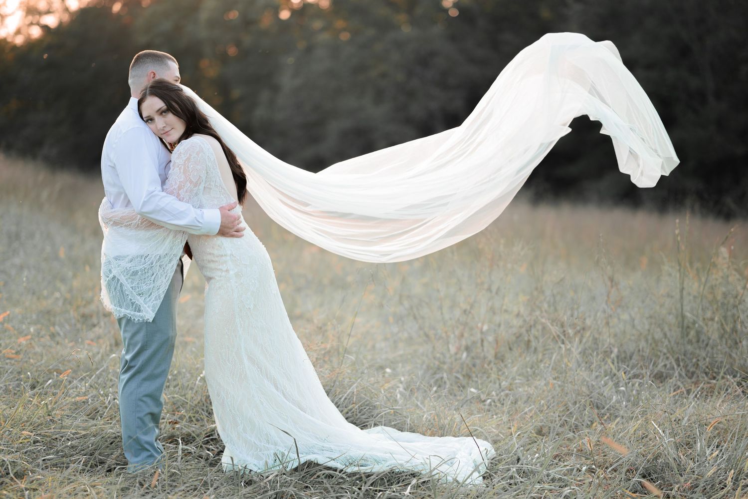A romantic wedding photo in a field at sunset with a flowing white veil caught in the breeze.