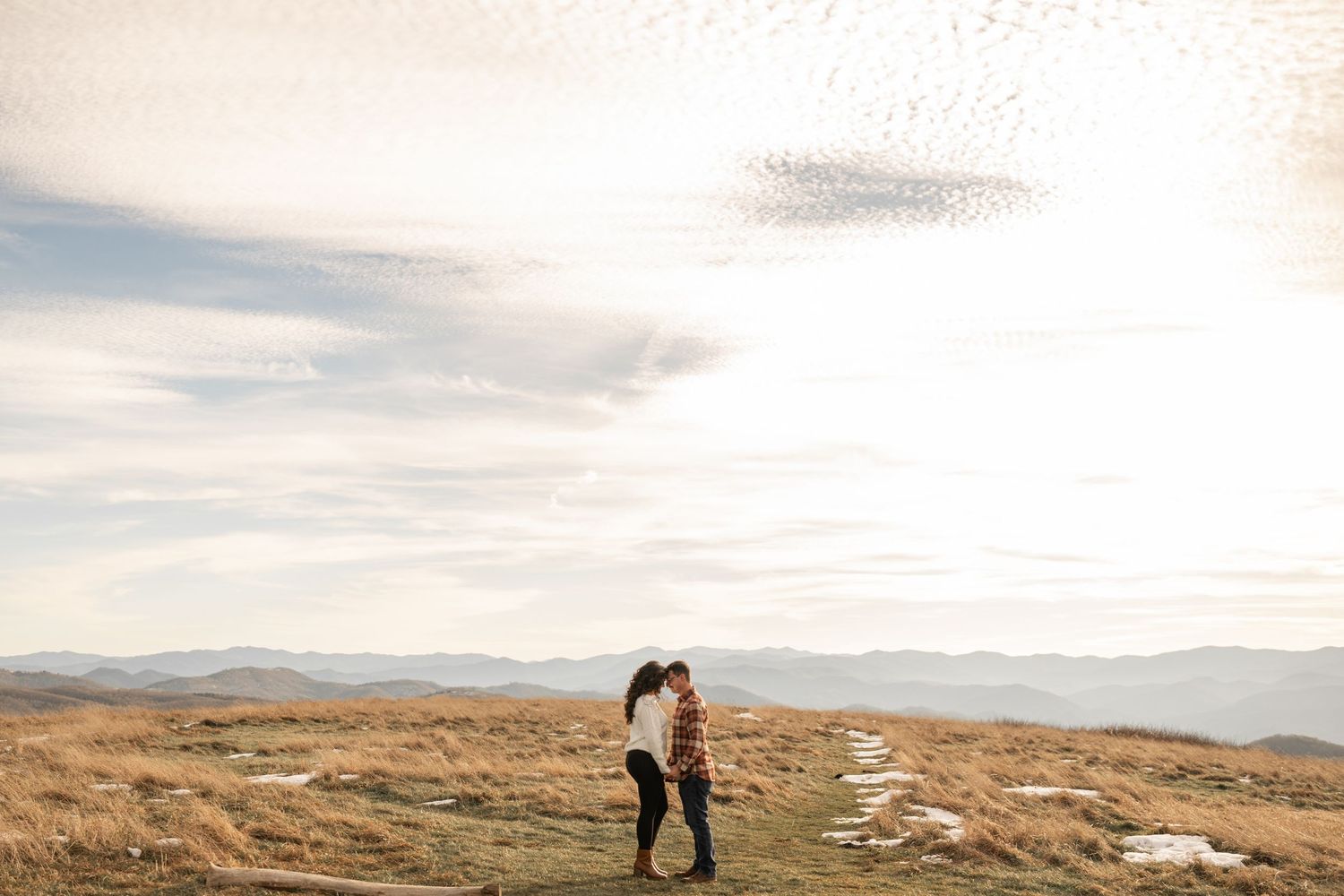 Couple embracing on hilltop against dramatic mountain landscape at sunset.