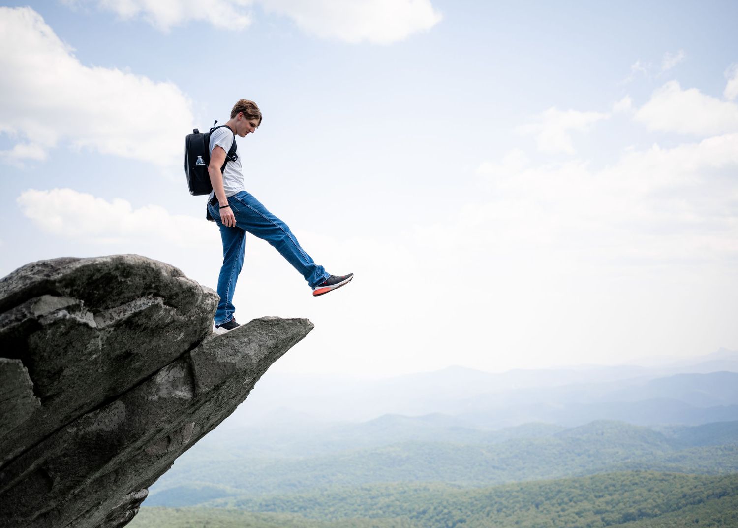 Hiker stands on edge of dramatic cliff overlook with mountain vista in background.
