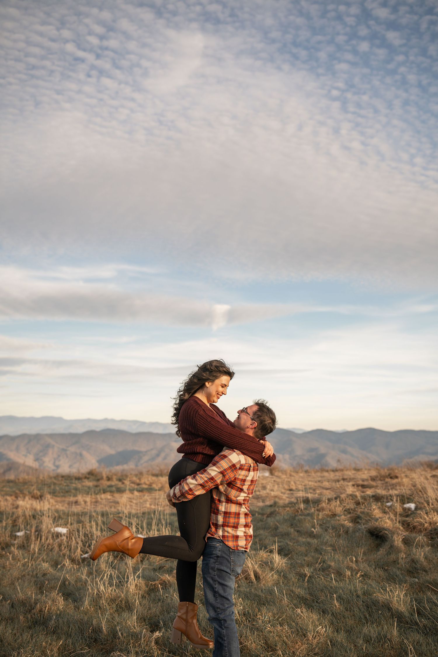 Playful couple spinning and embracing in mountain field under cloudy sky.