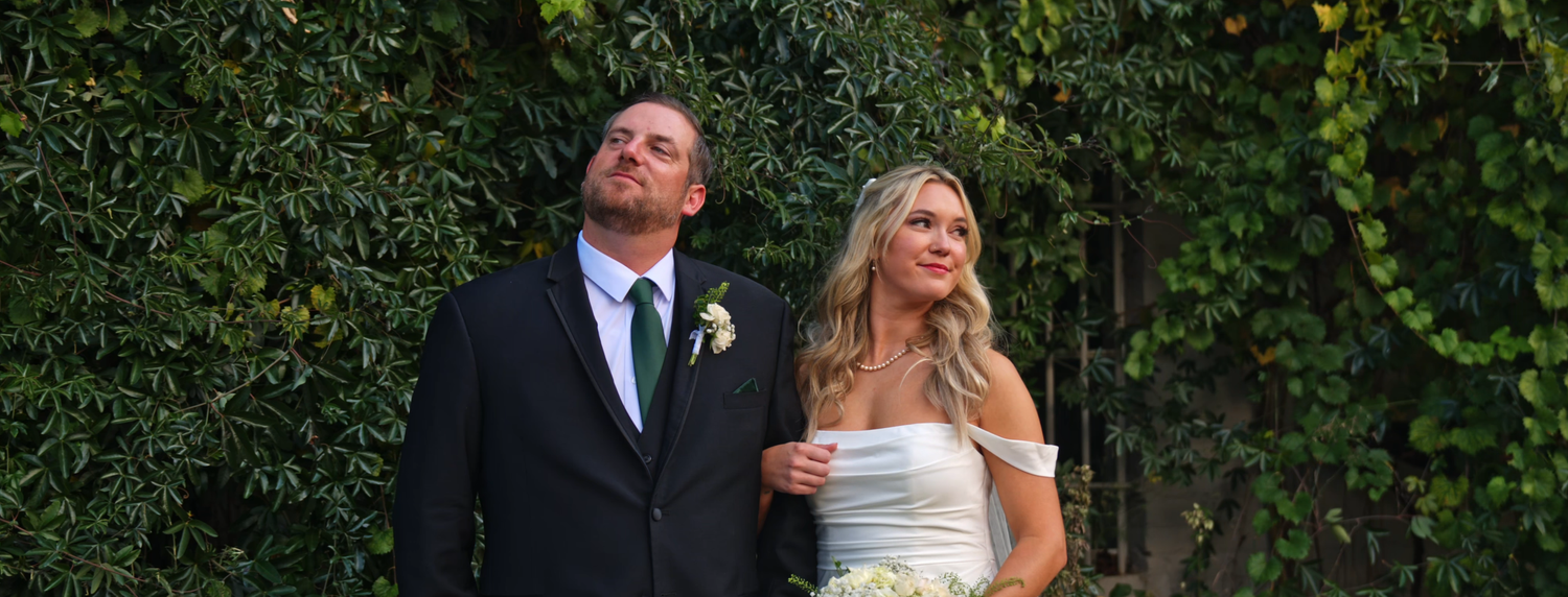 Bride and groom standing together outdoors in front of lush green ivy wall on their wedding day.