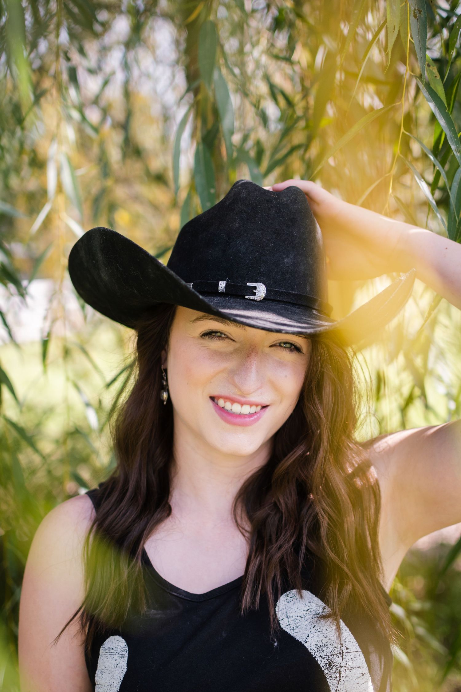 A portrait of someone in a black cowboy hat posing among willow branches in golden sunlight.