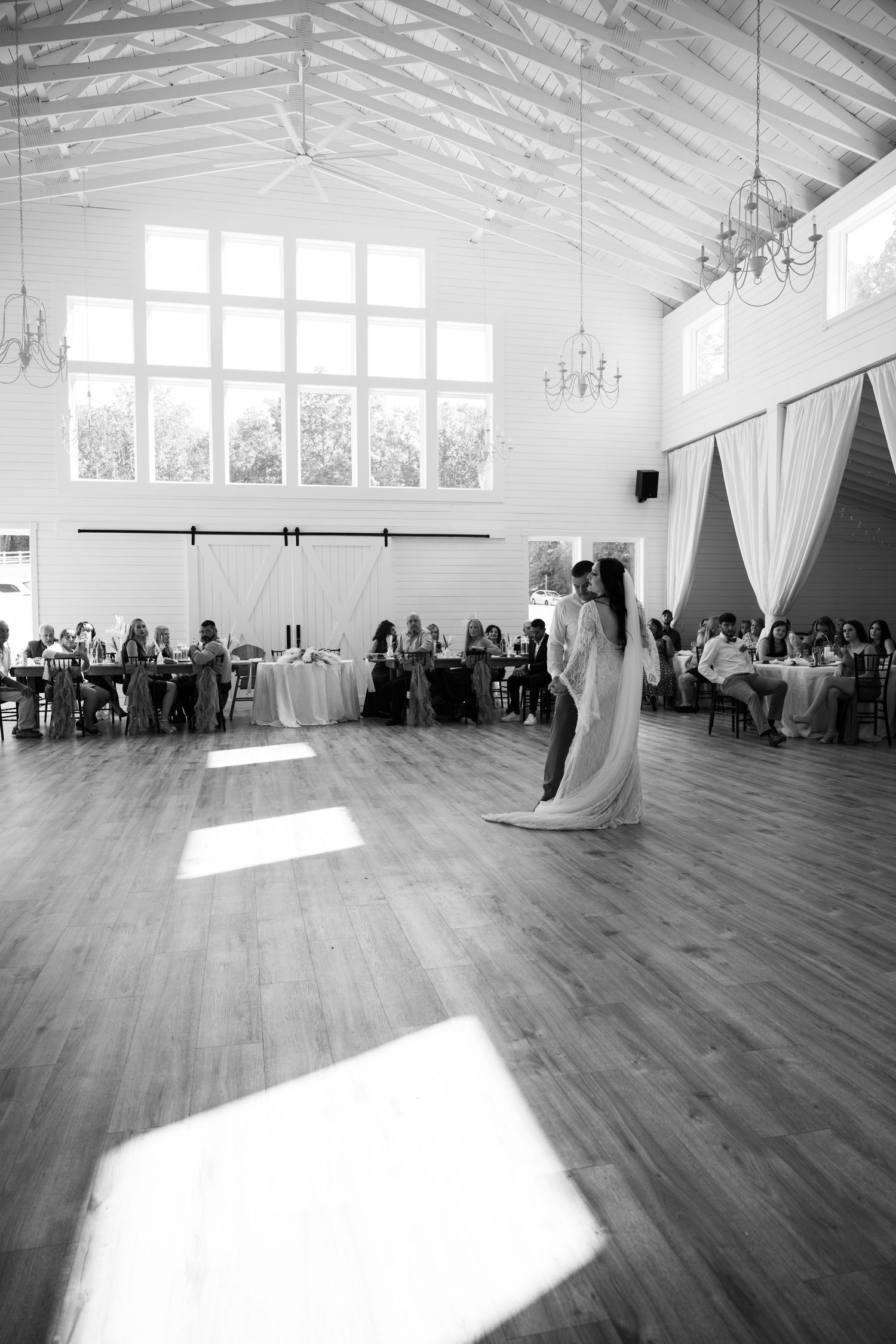 A spacious white reception hall with sunlight streaming through windows as a wedding dance unfolds.