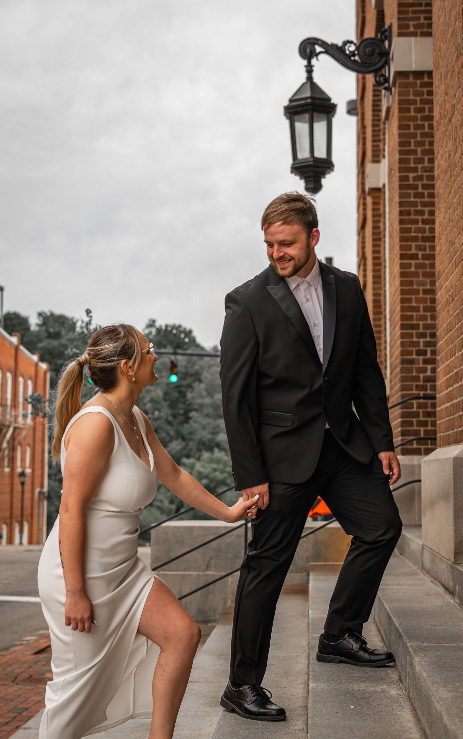 Playful couple in wedding attire posing on brick sidewalk with fun leg kicks.