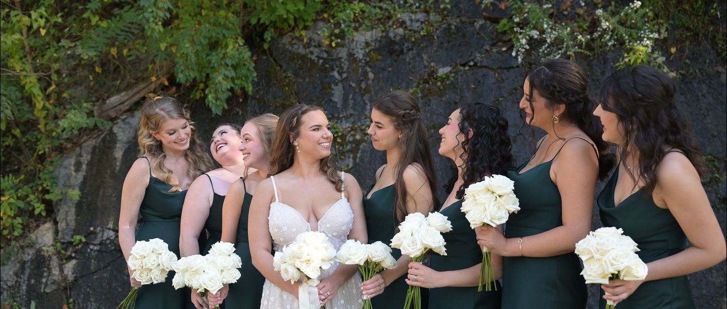 Bridesmaids in formal dresses holding white bouquets stand together outdoors.