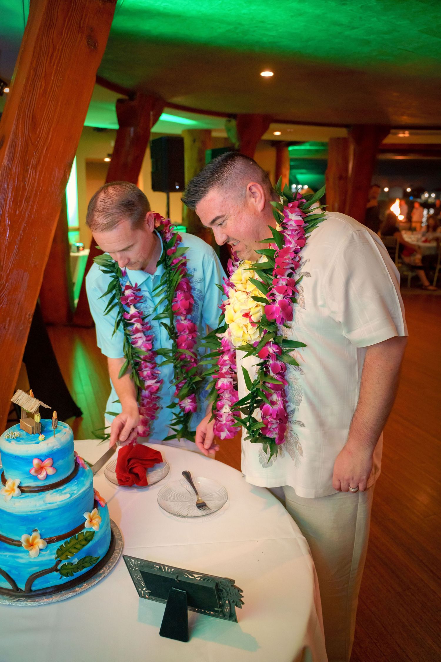 Two people in leis cutting a tropical-themed blue wedding cake with green lighting overhead.