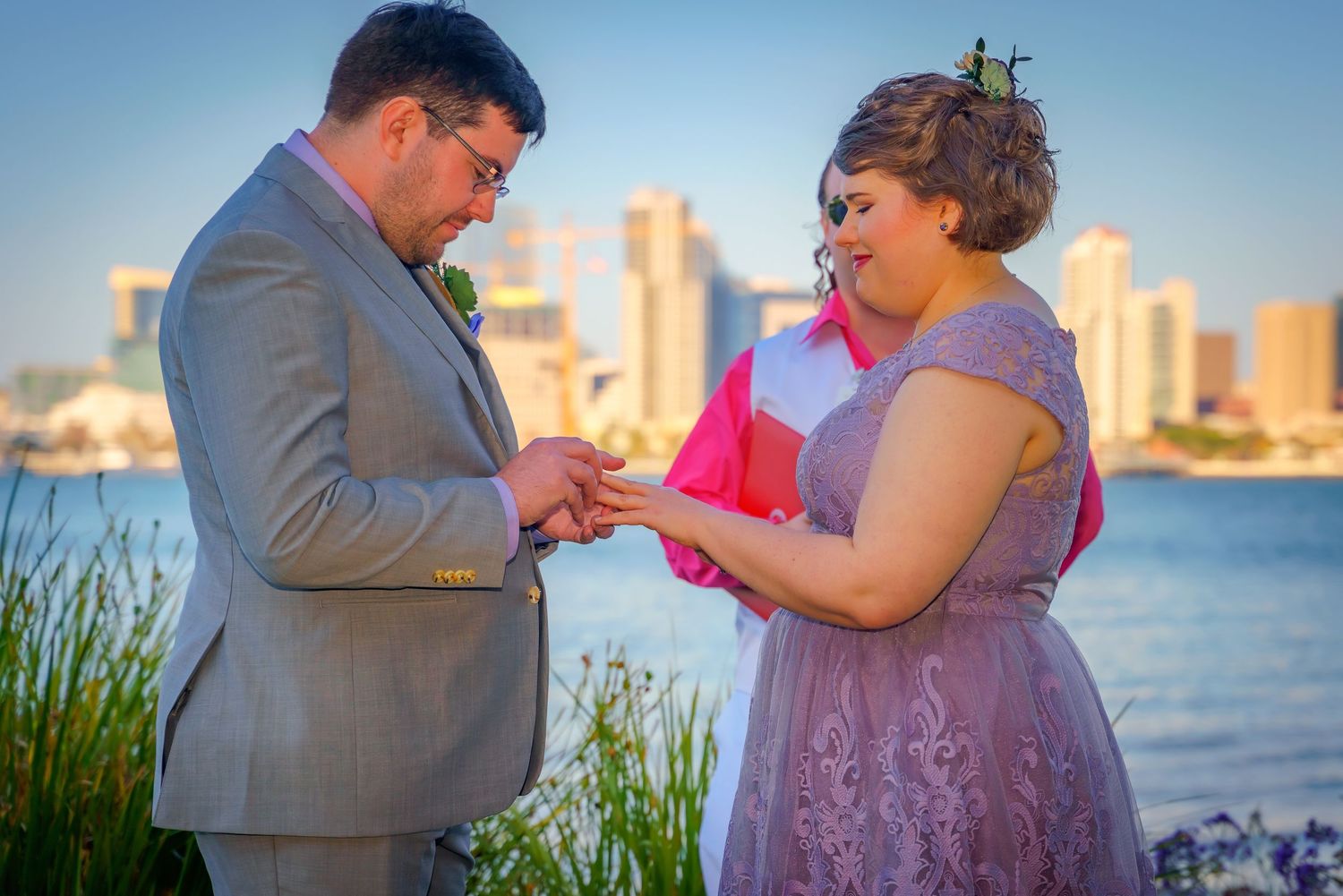 Couple exchanging rings during a waterfront ceremony with city skyline in background.