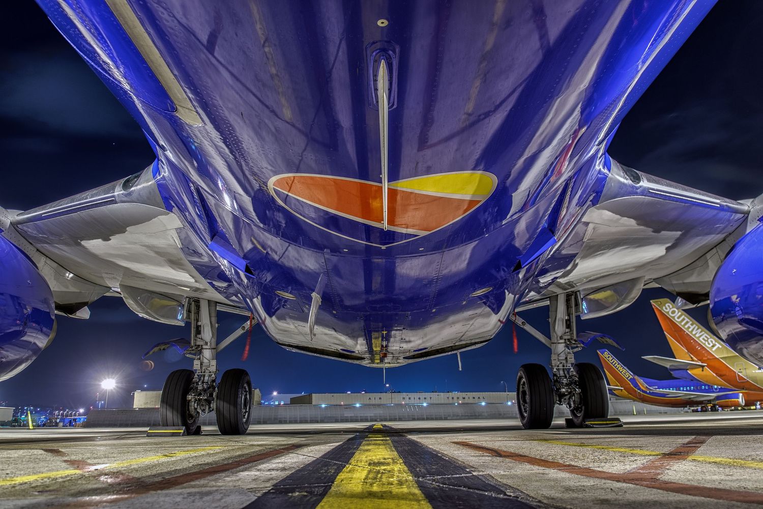 Under-wing perspective of commercial aircraft at night with blue and yellow lighting.