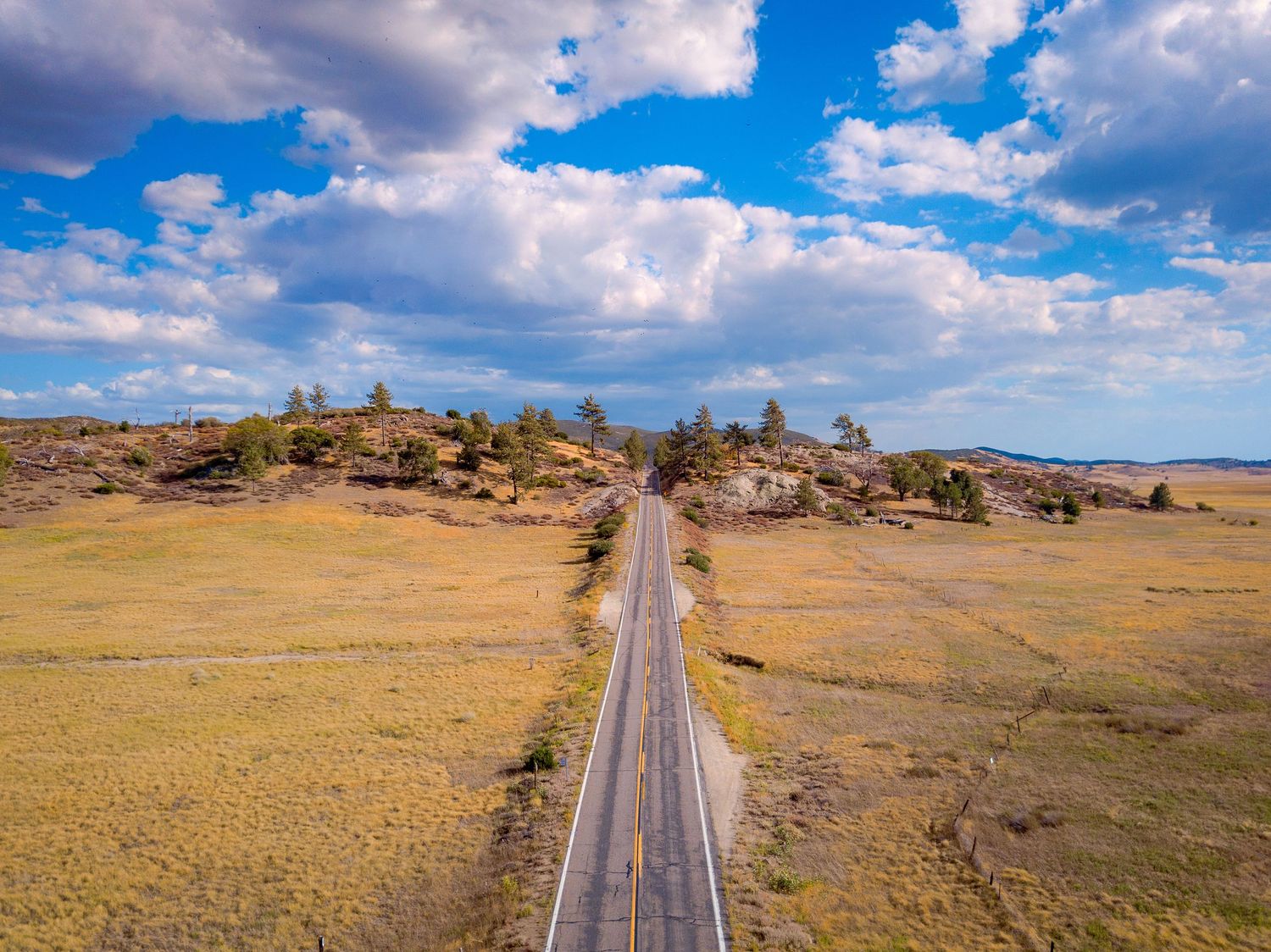 Aerial panorama of straight highway cutting through golden prairie grasslands under blue sky.