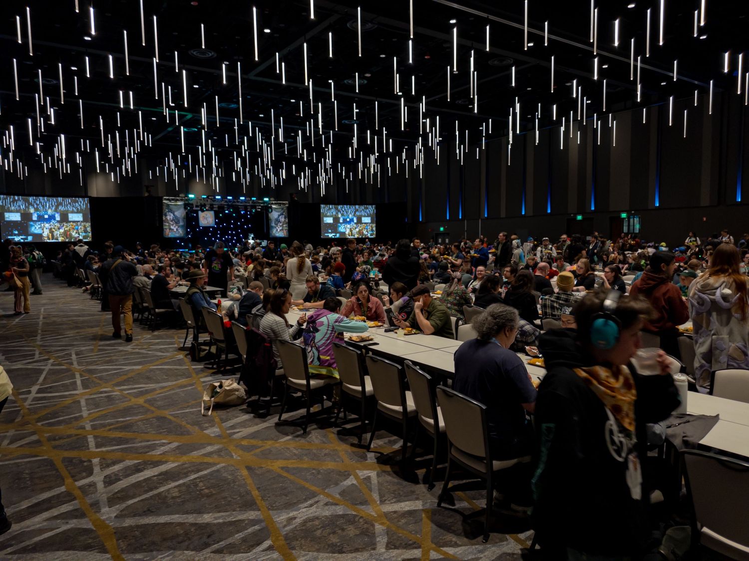 Large conference room filled with attendees seated at long tables under hanging lights during a professional event.