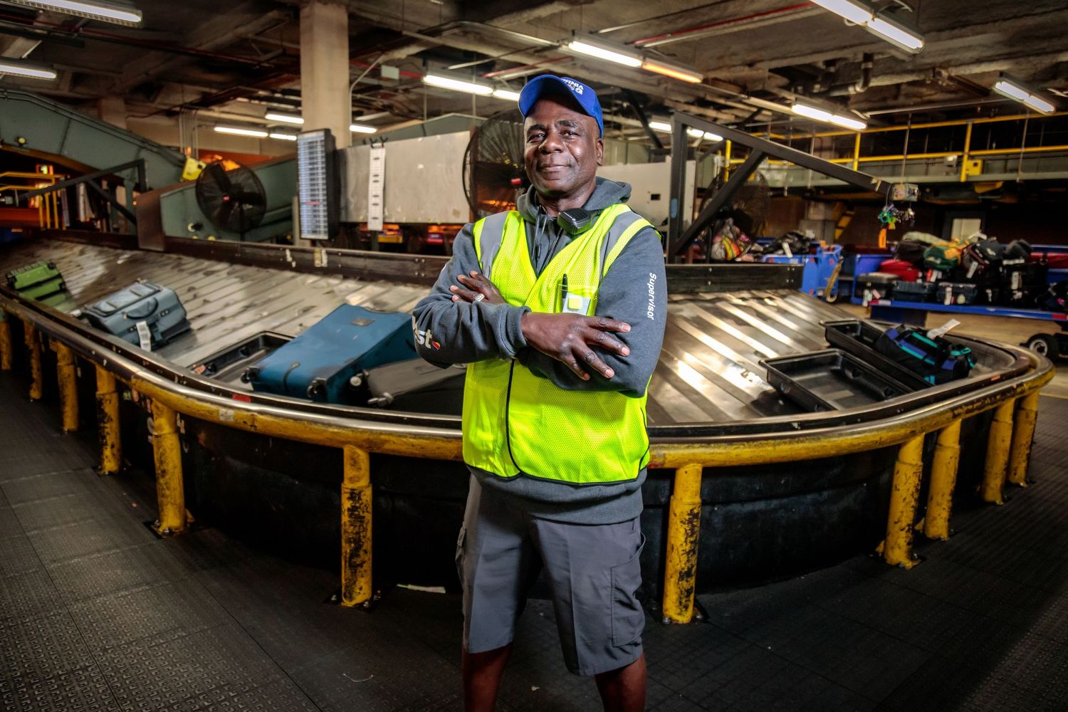 Workers in safety vests inspect luggage conveyor belts in airport baggage handling area.