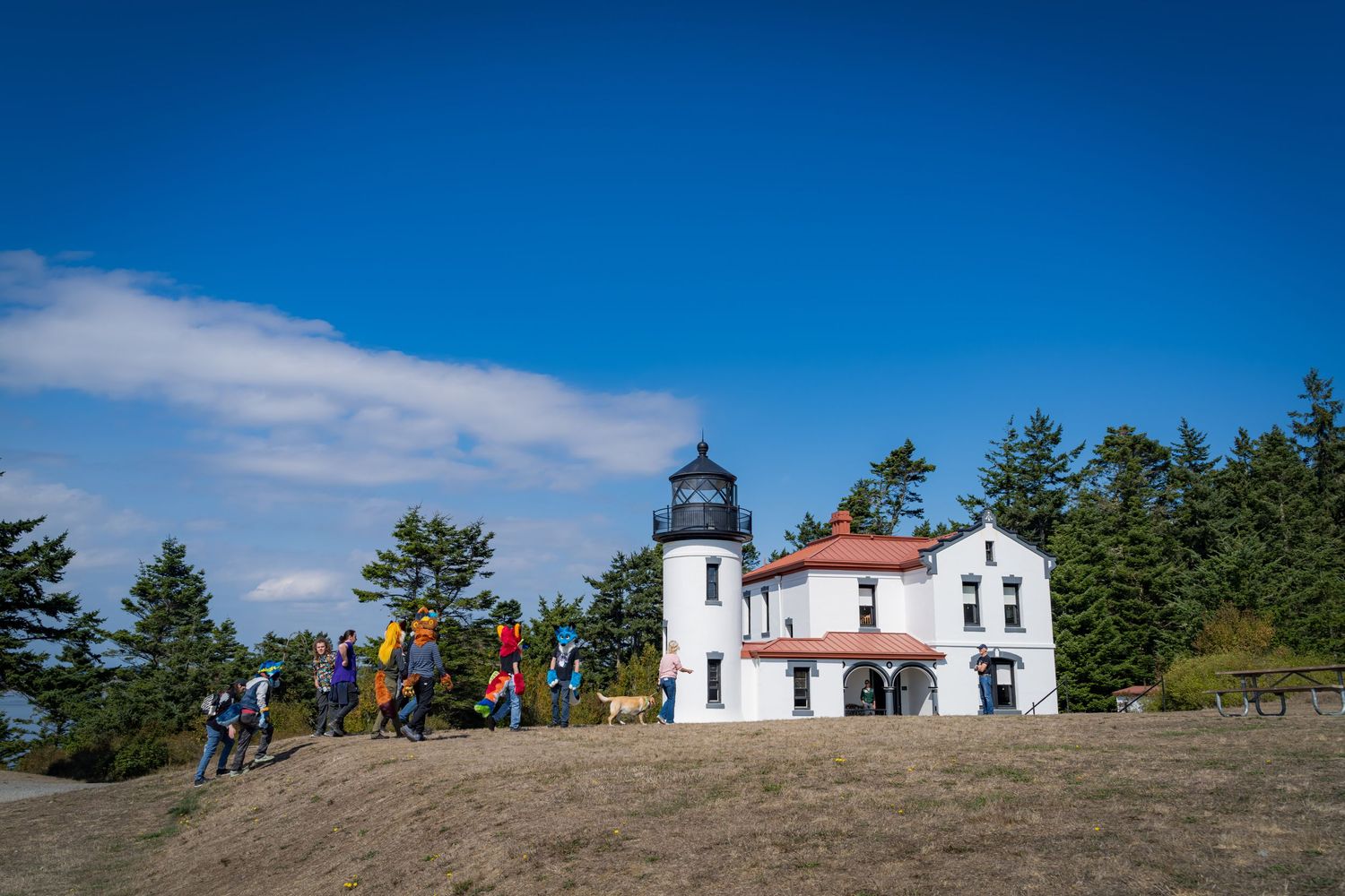 White lighthouse building under blue sky with people visible on hilltop.