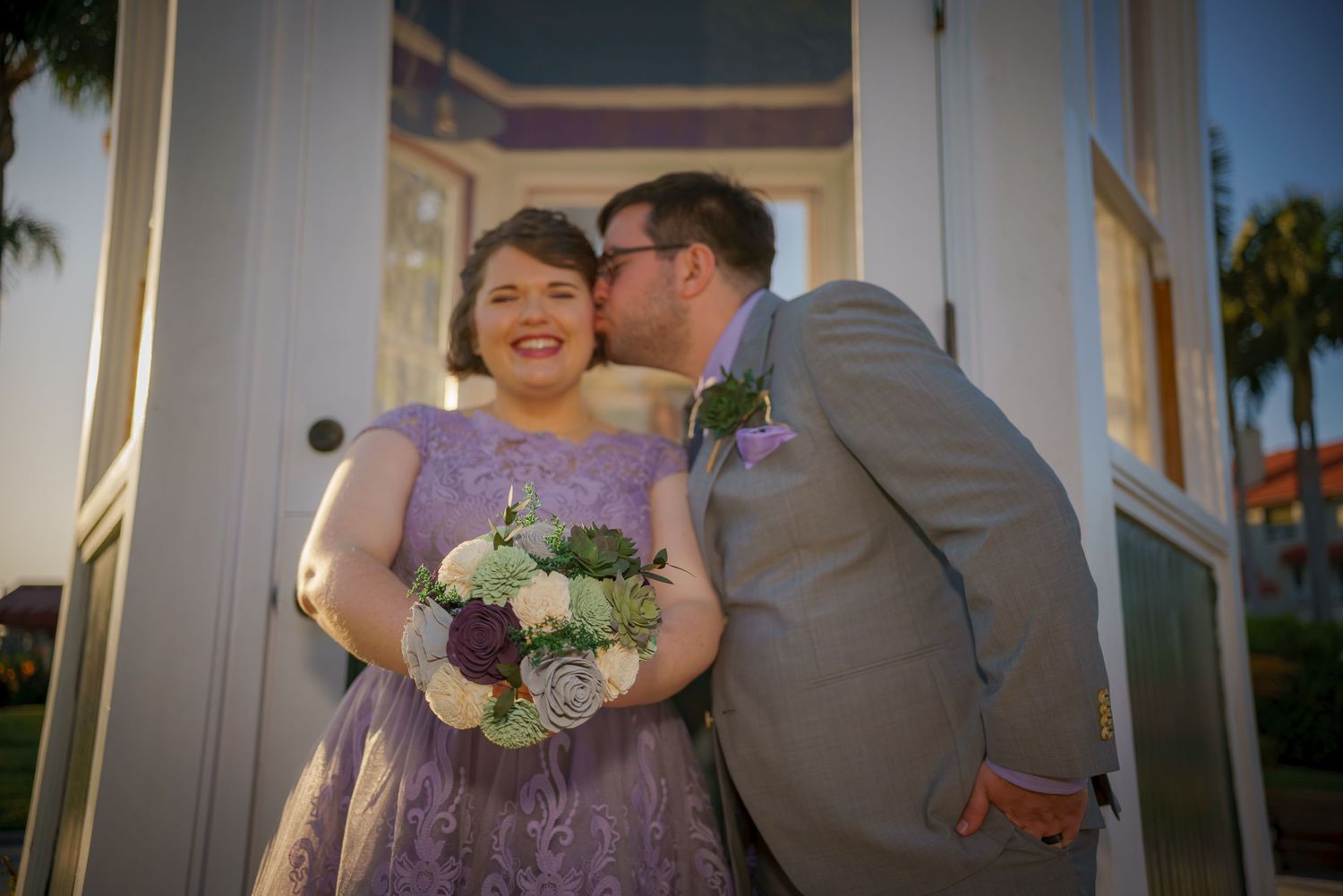 Wedding couple sharing intimate moment outside traditional white building with palm trees.