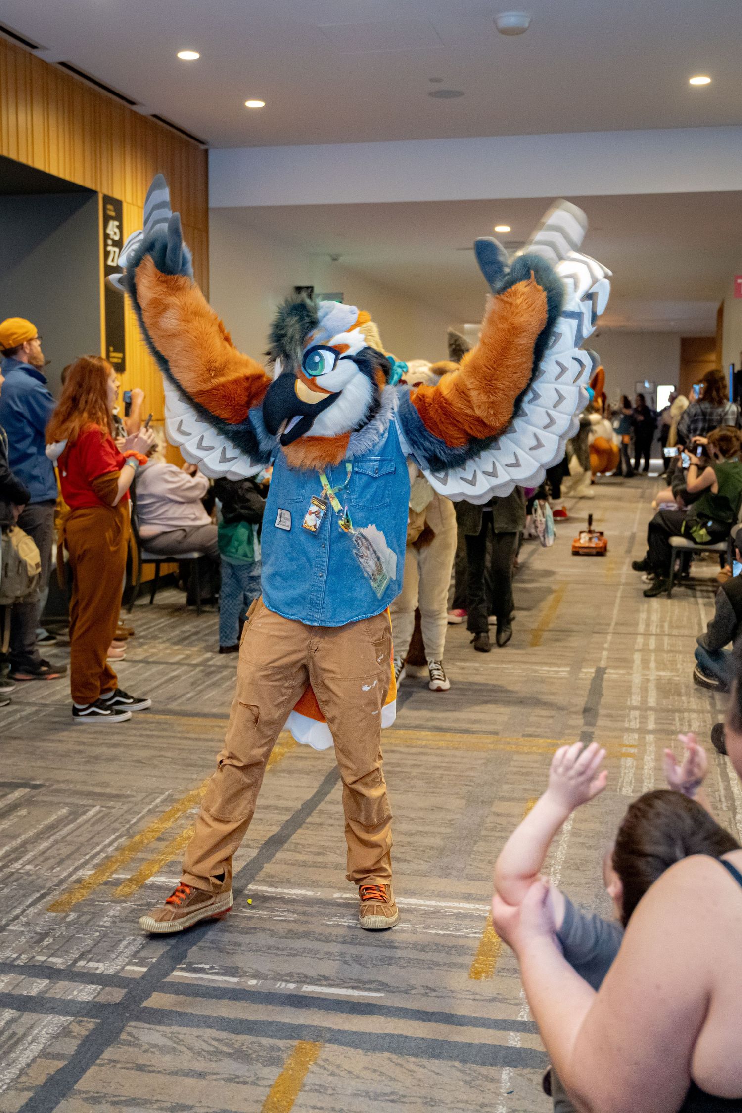 A furry mascot in brown and blue costume performs dance moves at an indoor convention space.