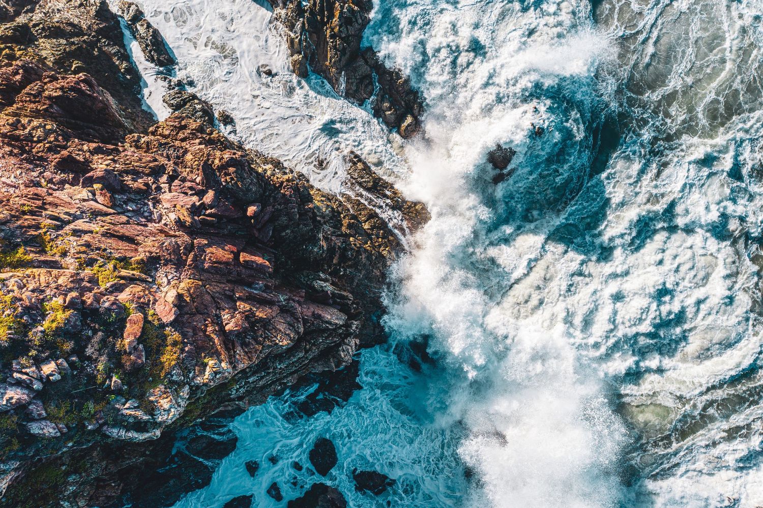 Dramatic aerial view of crashing ocean waves against rocky coastal cliffs with turquoise waters.