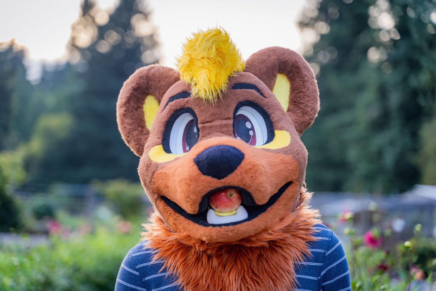 Brown fursuit head with large expressive eyes and fuzzy ears shown in close-up garden portrait.
